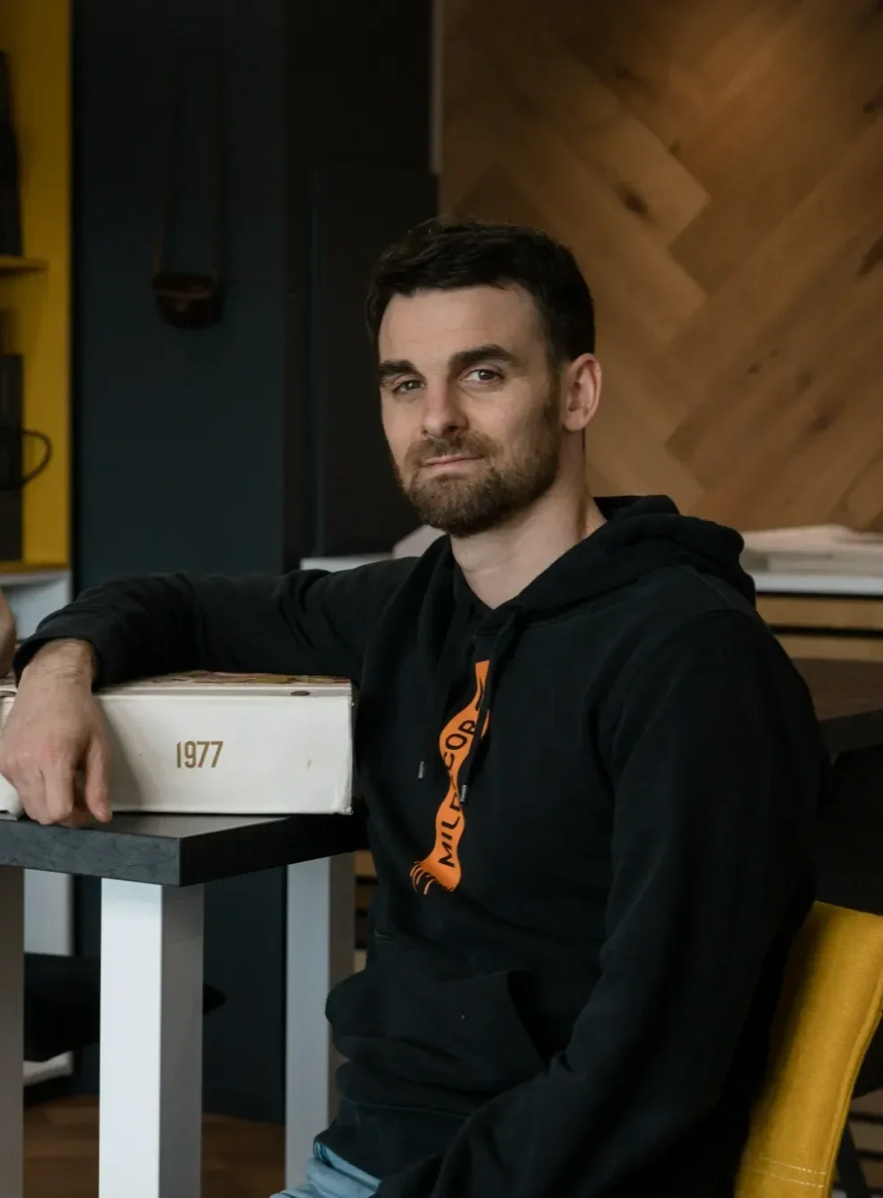 Un homme avec une barbe assis à une table dans une pièce avec un mur en bois, portant un sweat à capuche noir avec un logo orange, regardant vers la caméra.