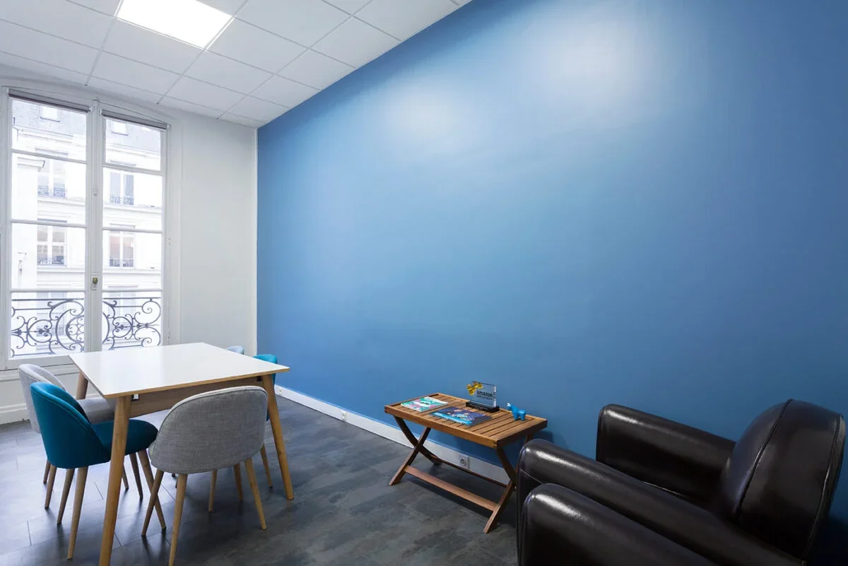 Une pièce avec un mur bleu, une table blanche avec quatre chaises colorées, deux fauteuils en cuir noir, une petite table en bois, et une grande fenêtre laissant entrer la lumière naturelle.