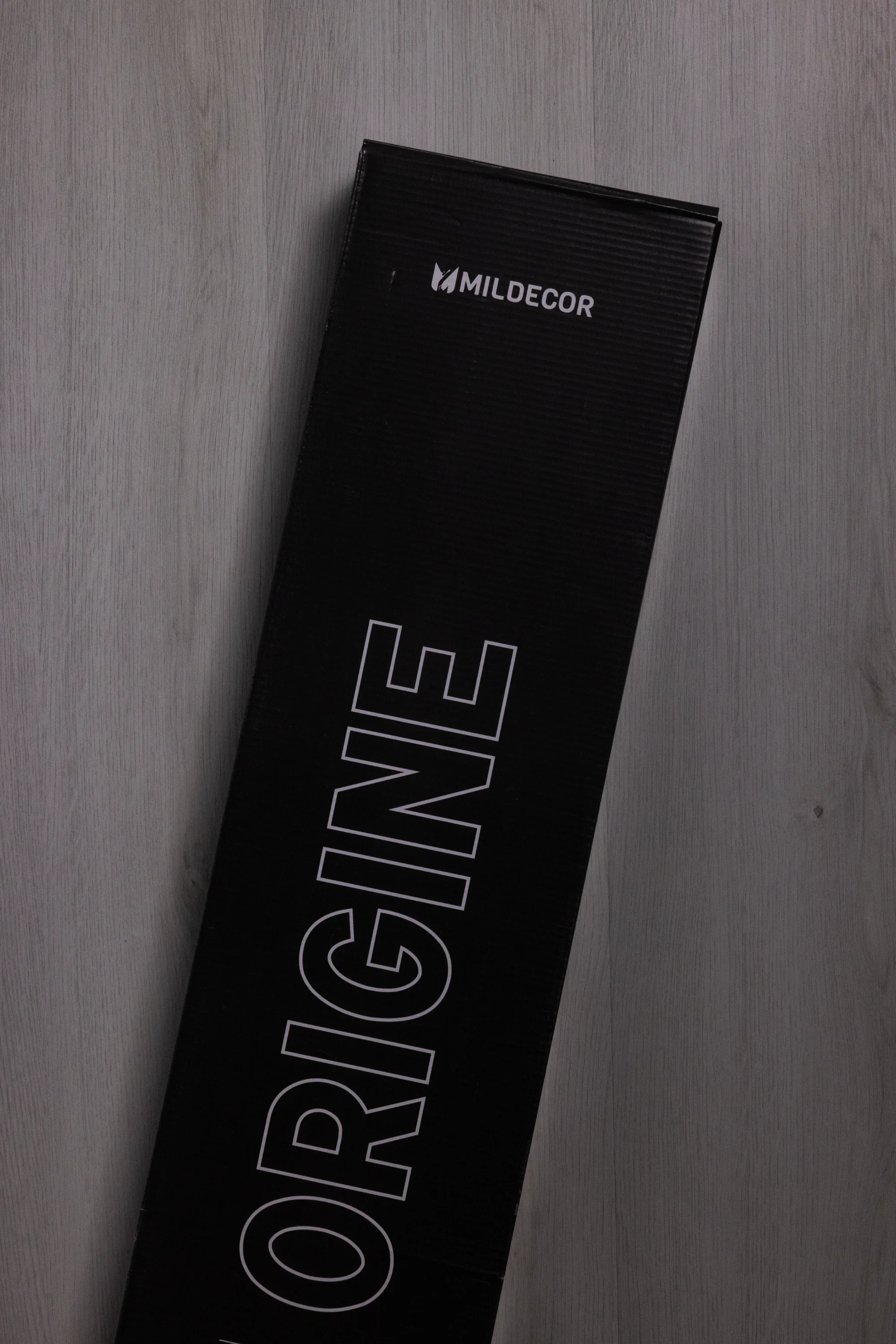Boîte noire portant le nom "ORIGININE" avec la marque Mildecor, placée à un angle sur un sol en bois clair.