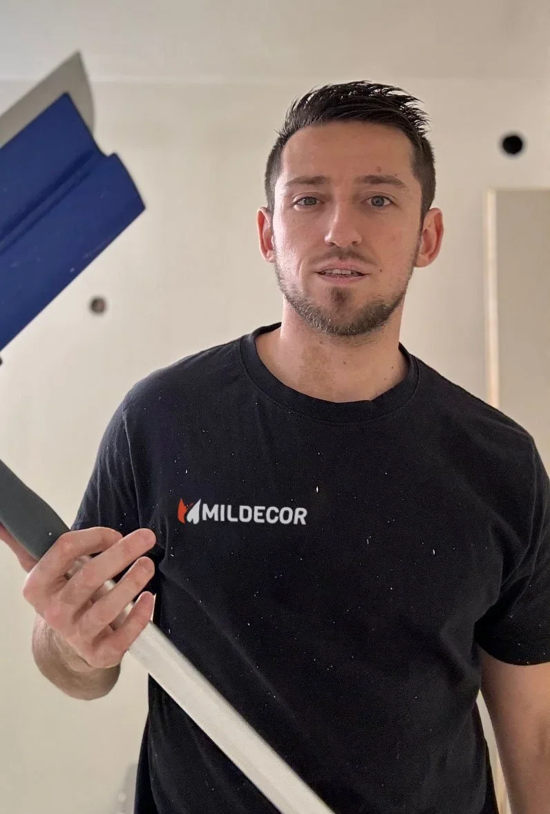 Un homme portant un t-shirt noir avec le logo 'MILDECOR' se tient devant un miroir, tenant un outil de peinture ou un rouleau à peinture.