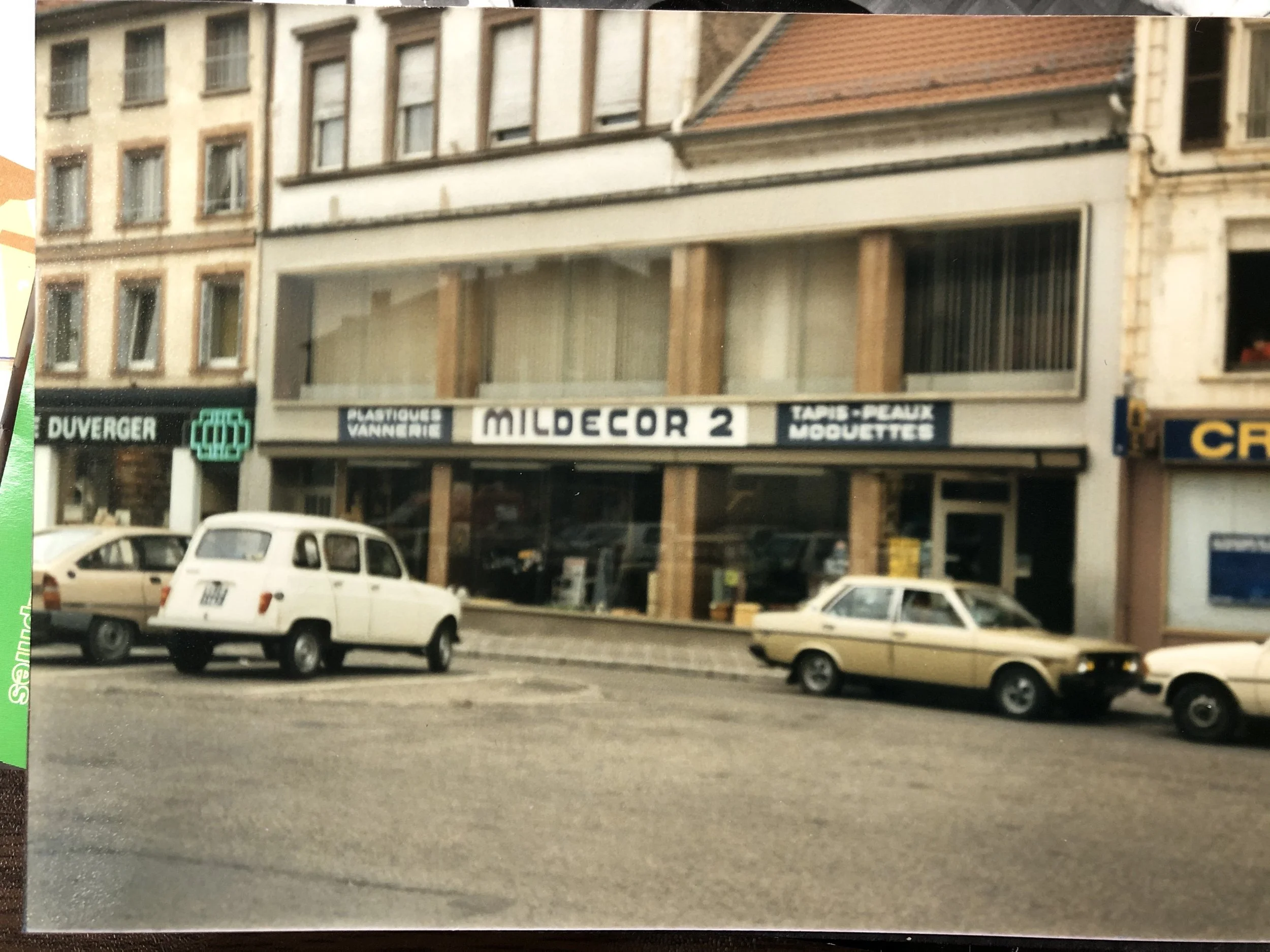 Une rue urbaine avec plusieurs voitures anciennes garées devant un bâtiment commerce avec une enseigne "MILDECOR 2" et autres magasins. En arrière-plan, des bâtiments à plusieurs étages et fenêtres.