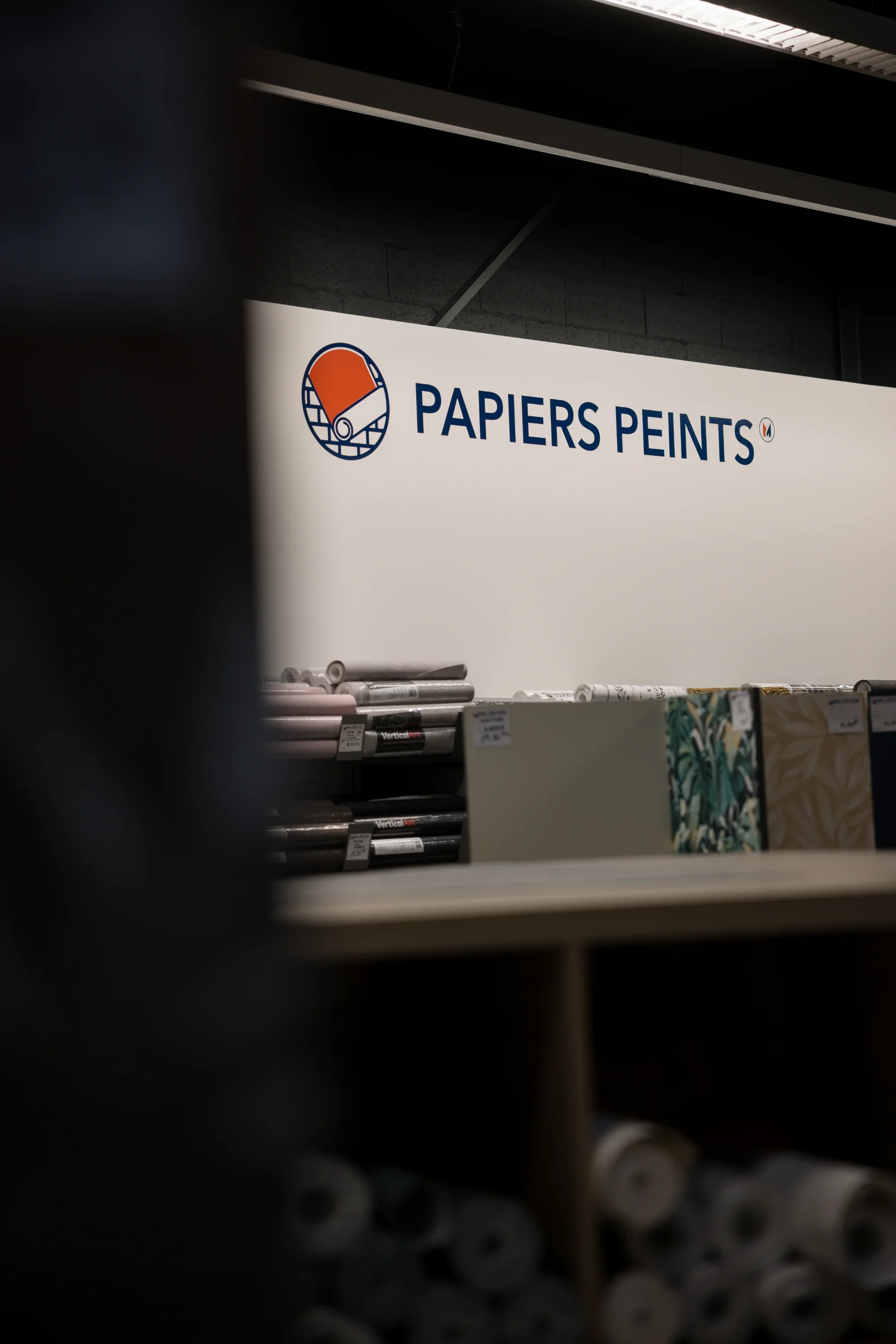 Une affiche dans un magasin de peinture affichant le logo et le nom 'Papiers Peints', avec des rouleaux de papier peint en dessous.