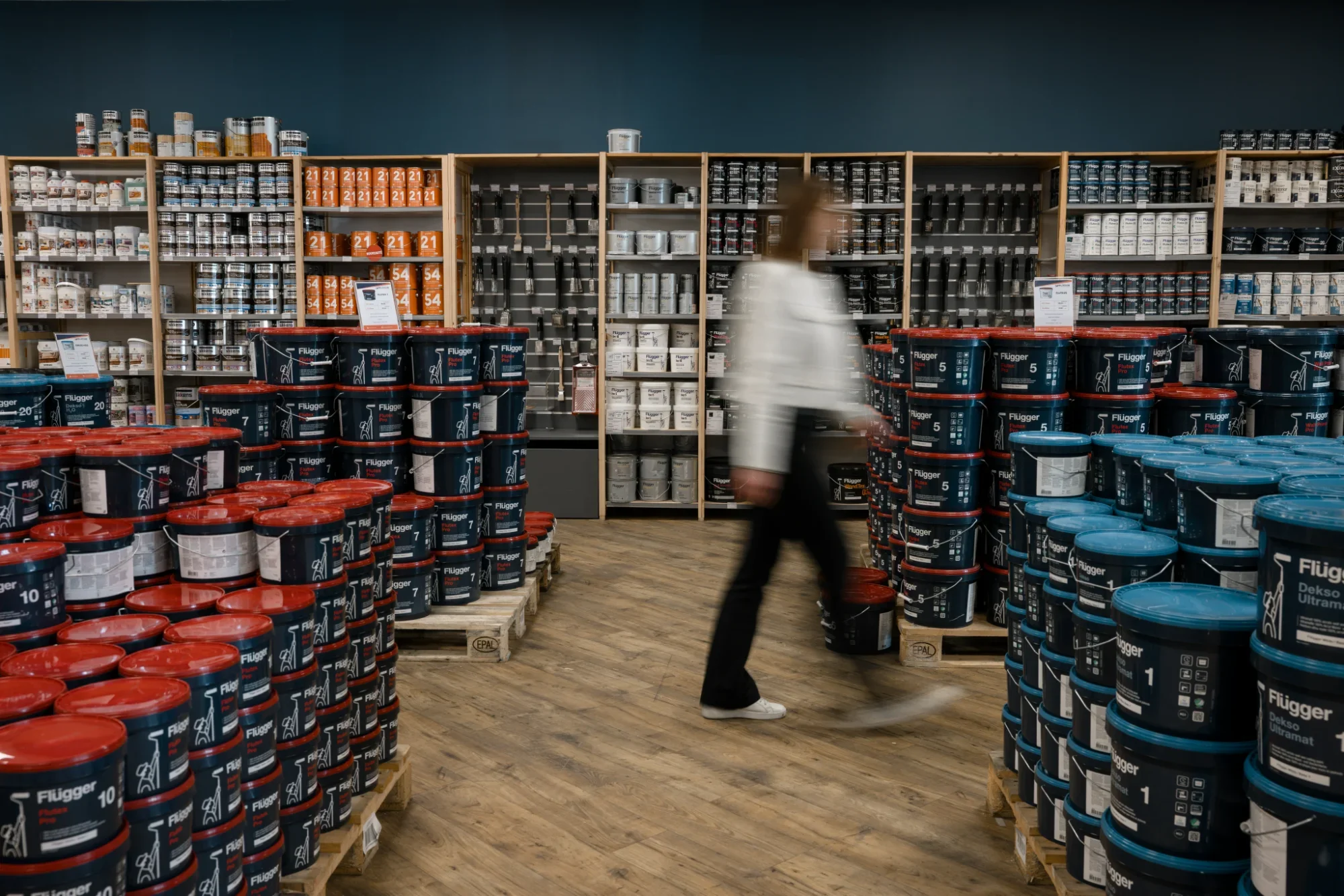 Une personne marche dans un magasin spécialisé en peinture, entourée de seaux de peinture de différentes tailles et couleurs, avec des rayons remplis de produits en arrière-plan.