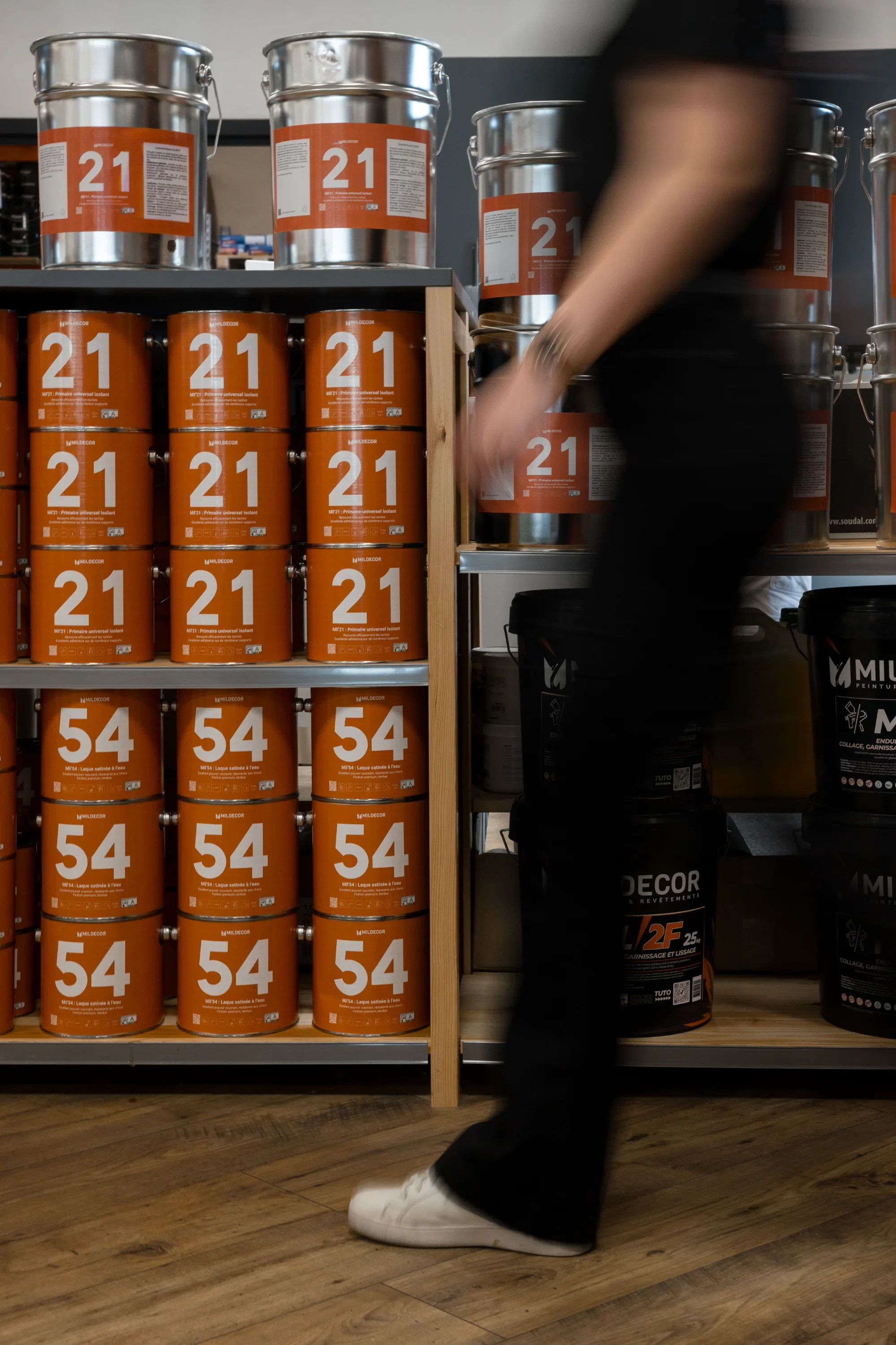 Près de rayons de peintures en endroit de magasin, une personne portant un pantalon noir et une chemise noire est en train de faire des achats.