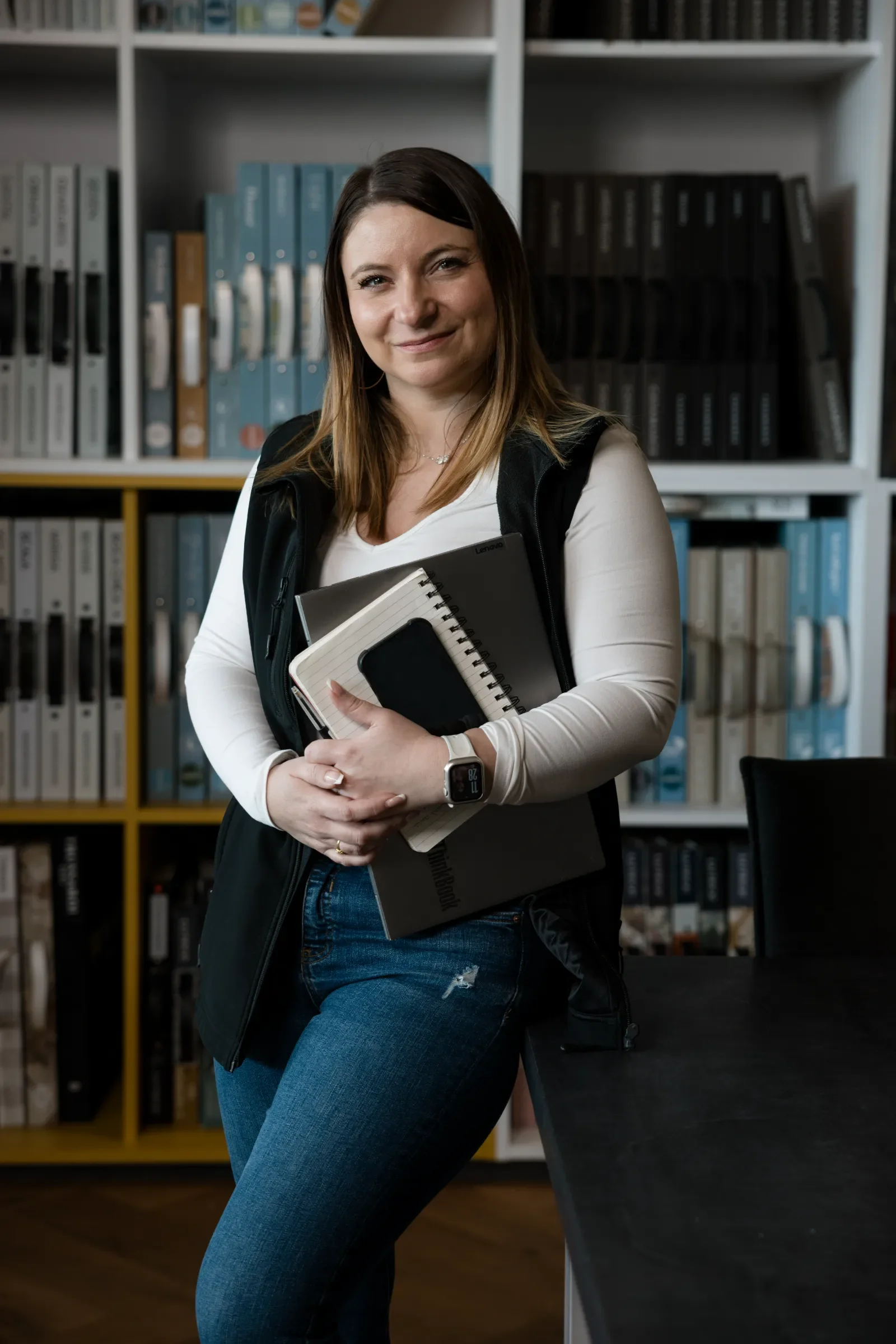 Une femme souriante debout dans une bibliothèque ou un bureau, tenant un carnet, un ordinateur portable et un téléphone, vêtue d'un sweat noir et d'un jean déchiré.