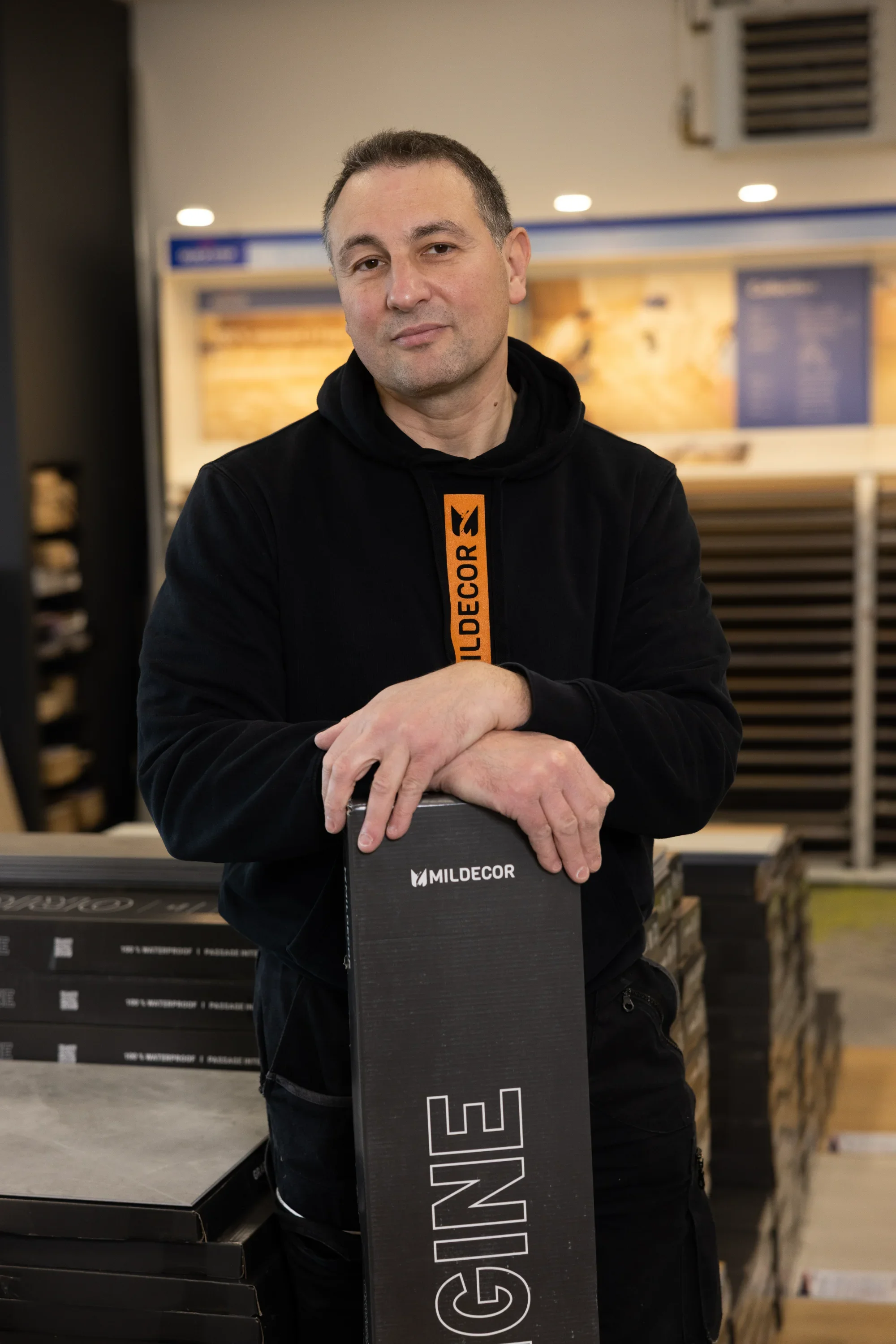 Un homme souriant, portant un hoodie noir avec la chaîne orange 'MILDDECOR', pose avec une boîte noire avec le logo 'MILDDECOR' dans un magasin de décoration ou de bricolage.
