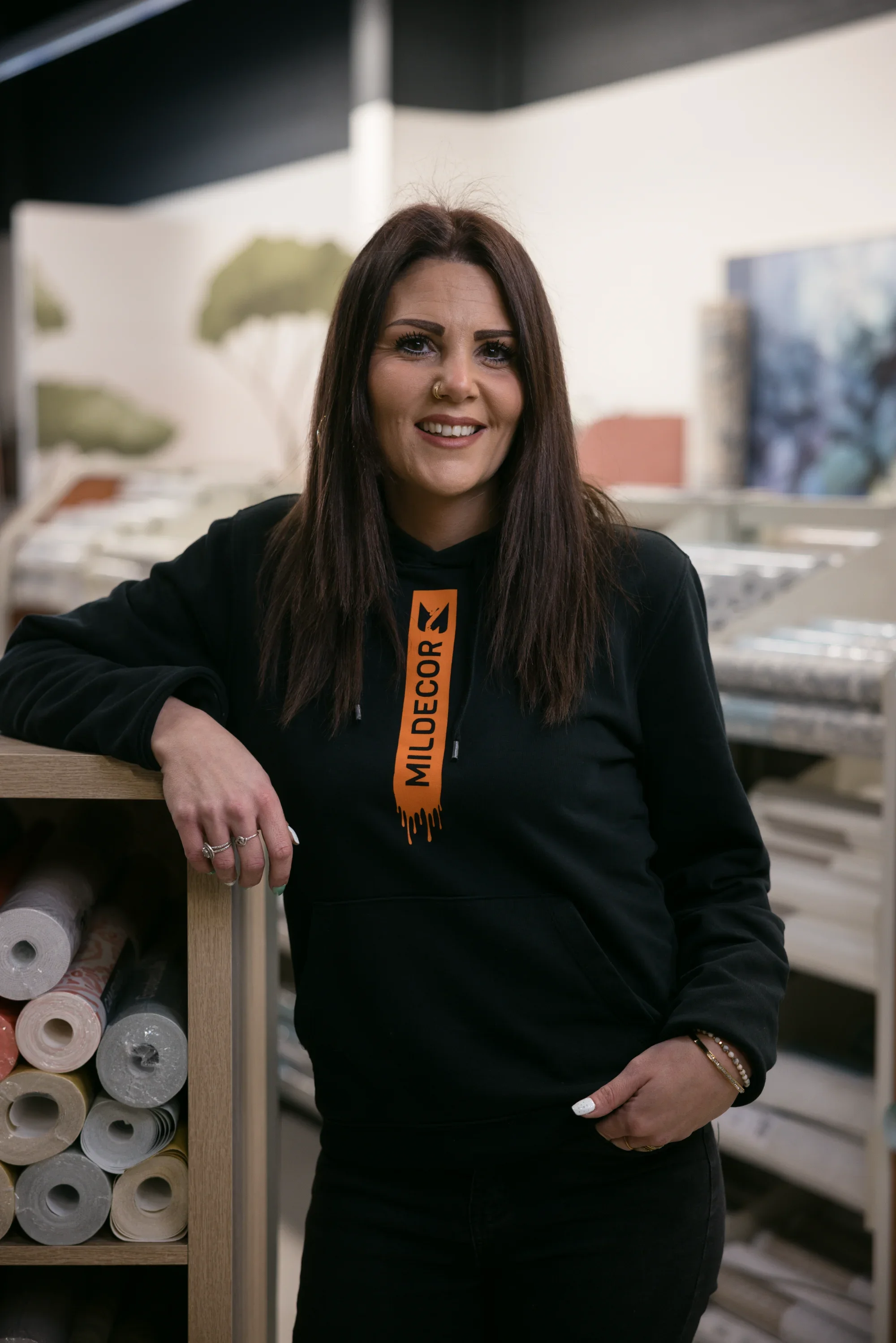 Femme souriante portant un sweat-shirt noir avec le logo Mildecore, posant dans un magasin de décoration avec des rouleaux de papier peint sur une étagère à sa gauche et des articles de décoration en arrière-plan.