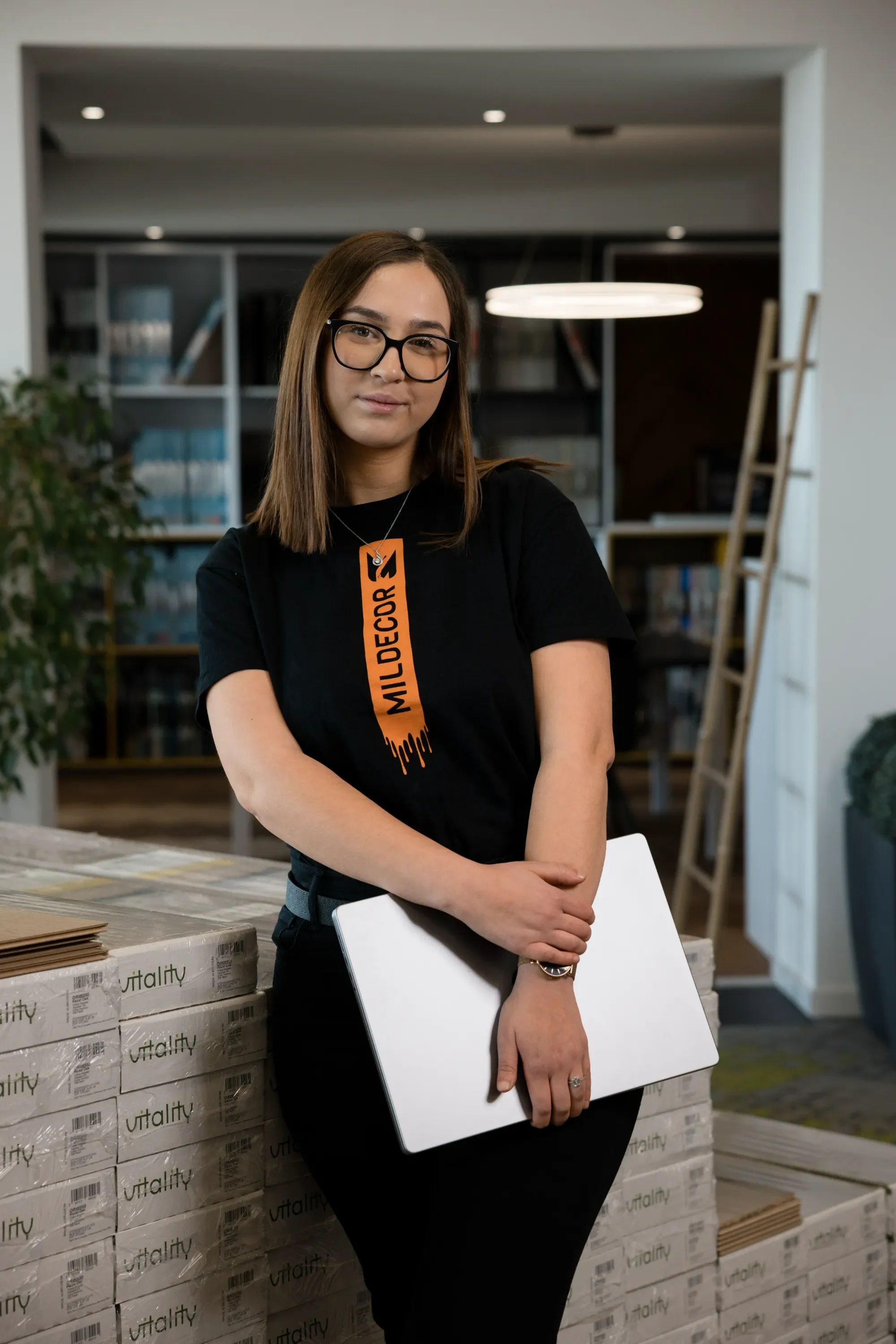 Jeune femme avec lunettes, portant un t-shirt noir avec une étiquette orange disant 'MILLDECORE', tenant un ordinateur portable blanc, se tenant devant des boîtes empilées avec le mot 'utality' écrit dessus dans un environnement intérieur moderne.