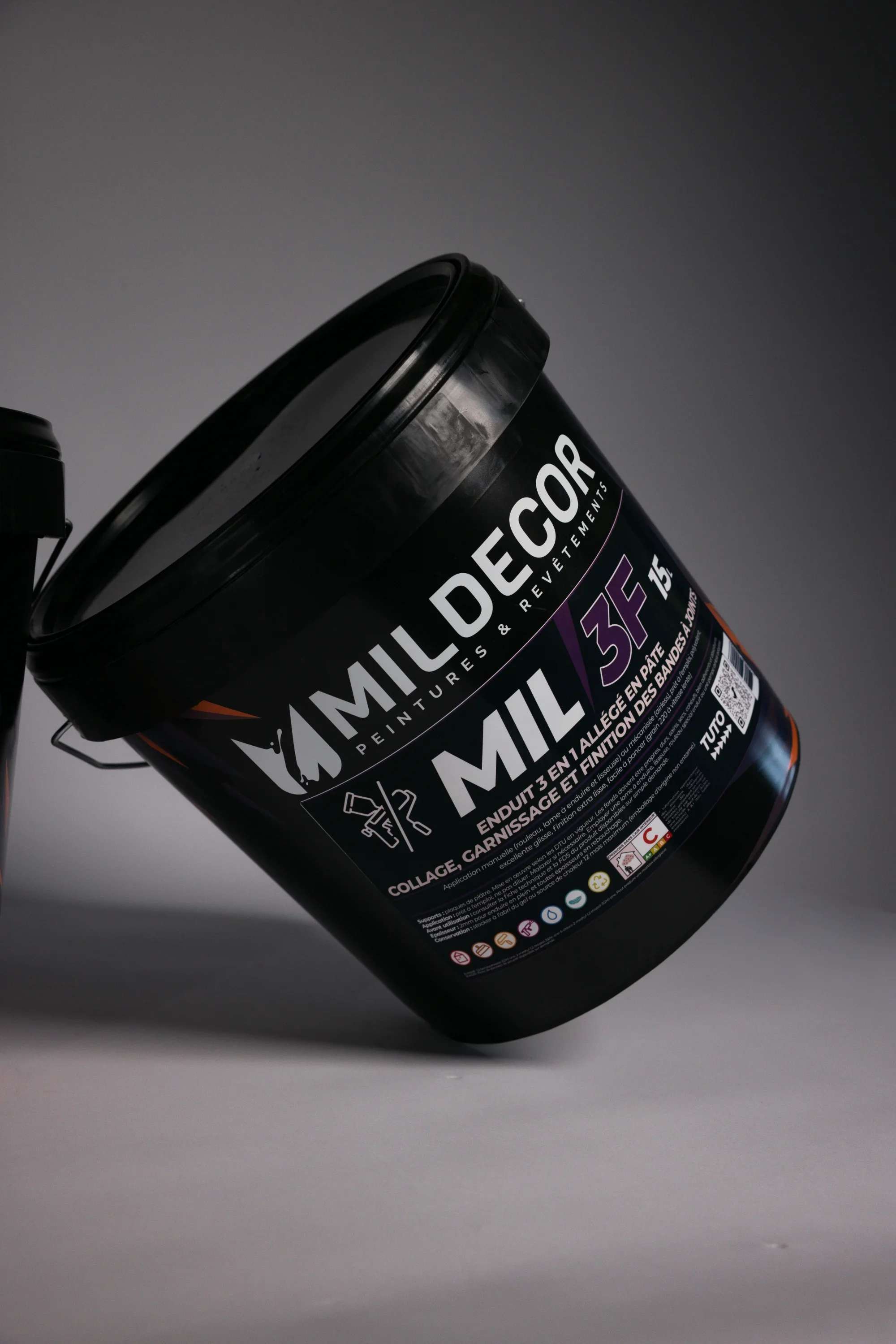 Un pot de peinture noir de la marque Mildecor, modèle MIL 35F, posé en diagonale contre un fond gris, avec étiquetage en français indiquant qu'il s'agit d'une peinture pour enduit et façade.