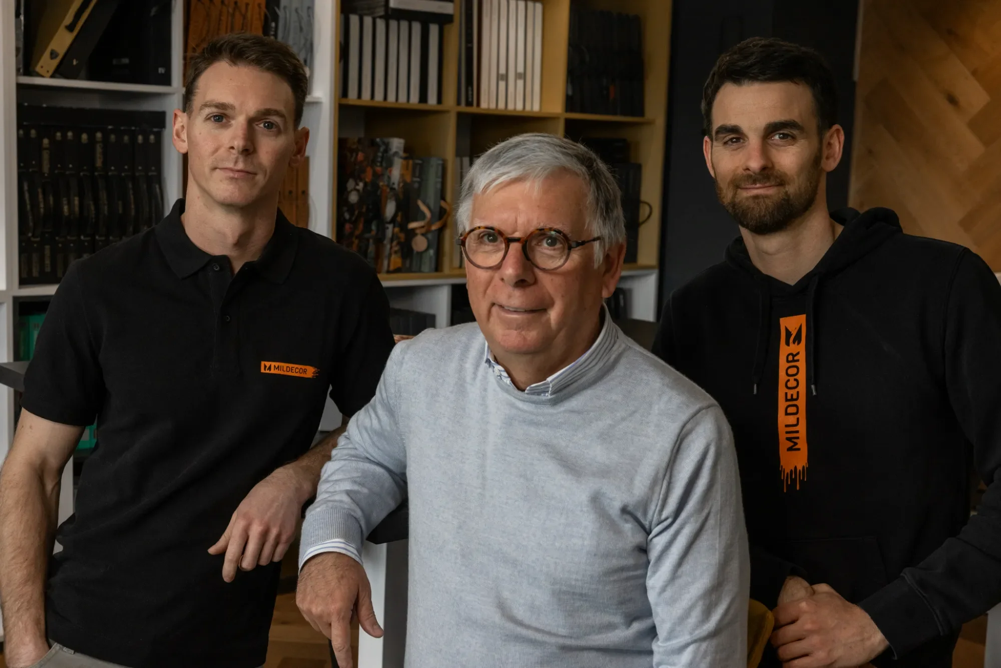 Trois hommes posent ensemble dans une pièce avec des étagères remplies de livres. L'homme au centre porte des lunettes et un pull gris, les deux autres portent des vêtements noirs avec des logos orange.