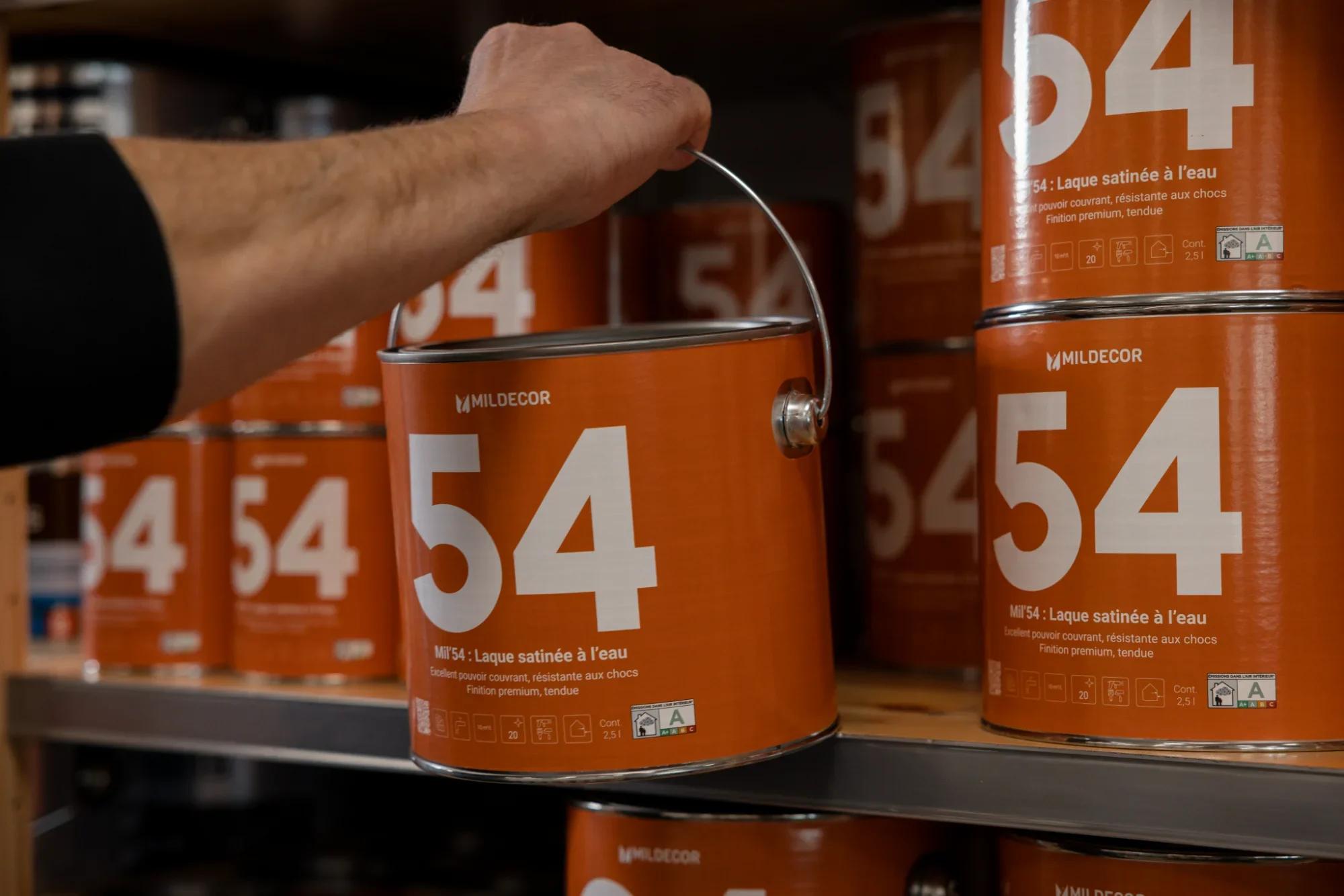 Un homme attrape un pot de peinture orange marqué du numéro 54 avec une poignée métallique, le pot est posé sur une étagère remplie d'autres pots de peinture du même modèle.