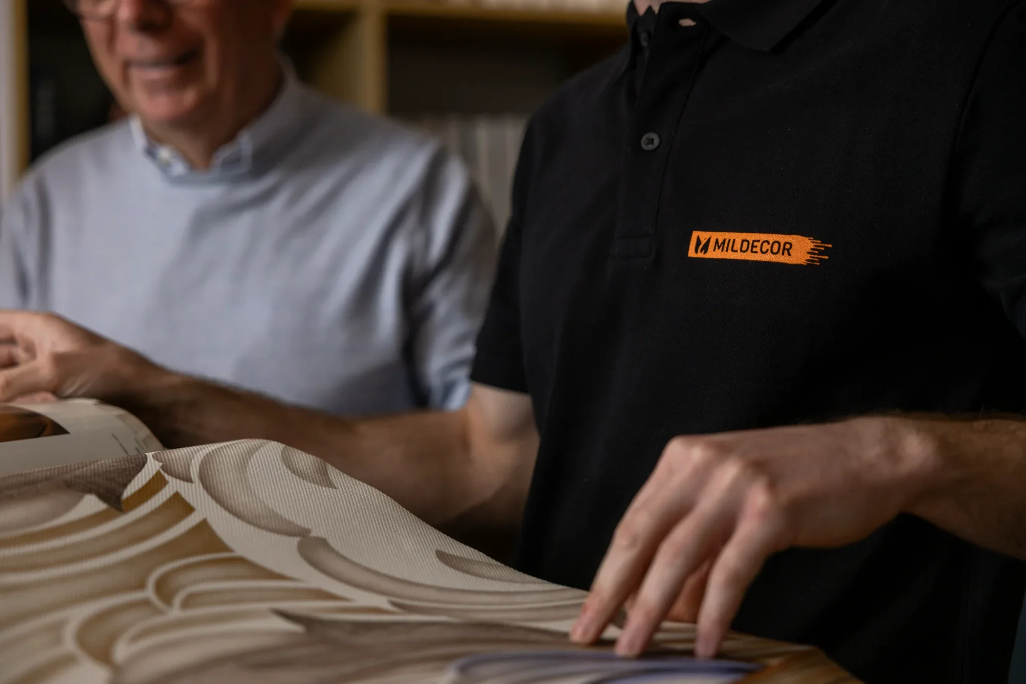 Deux hommes regardant un échantillon de tissu ou de papier avec un fond de bibliothèque, l'un portant un polo noir avec un logo orange, l'autre un chemisier gris clair.