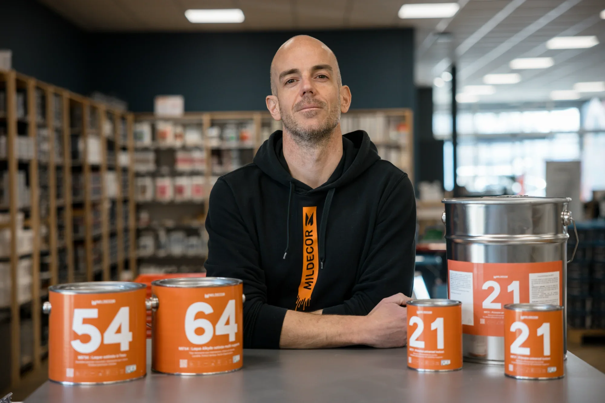 Un homme assis à une table avec plusieurs pots de peinture orange numérotés 54, 64, 21, 21, dans un magasin ou un atelier. Il porte un sweat à capuche noir avec une étiquette orange mentionnant 'MILDECORE'.