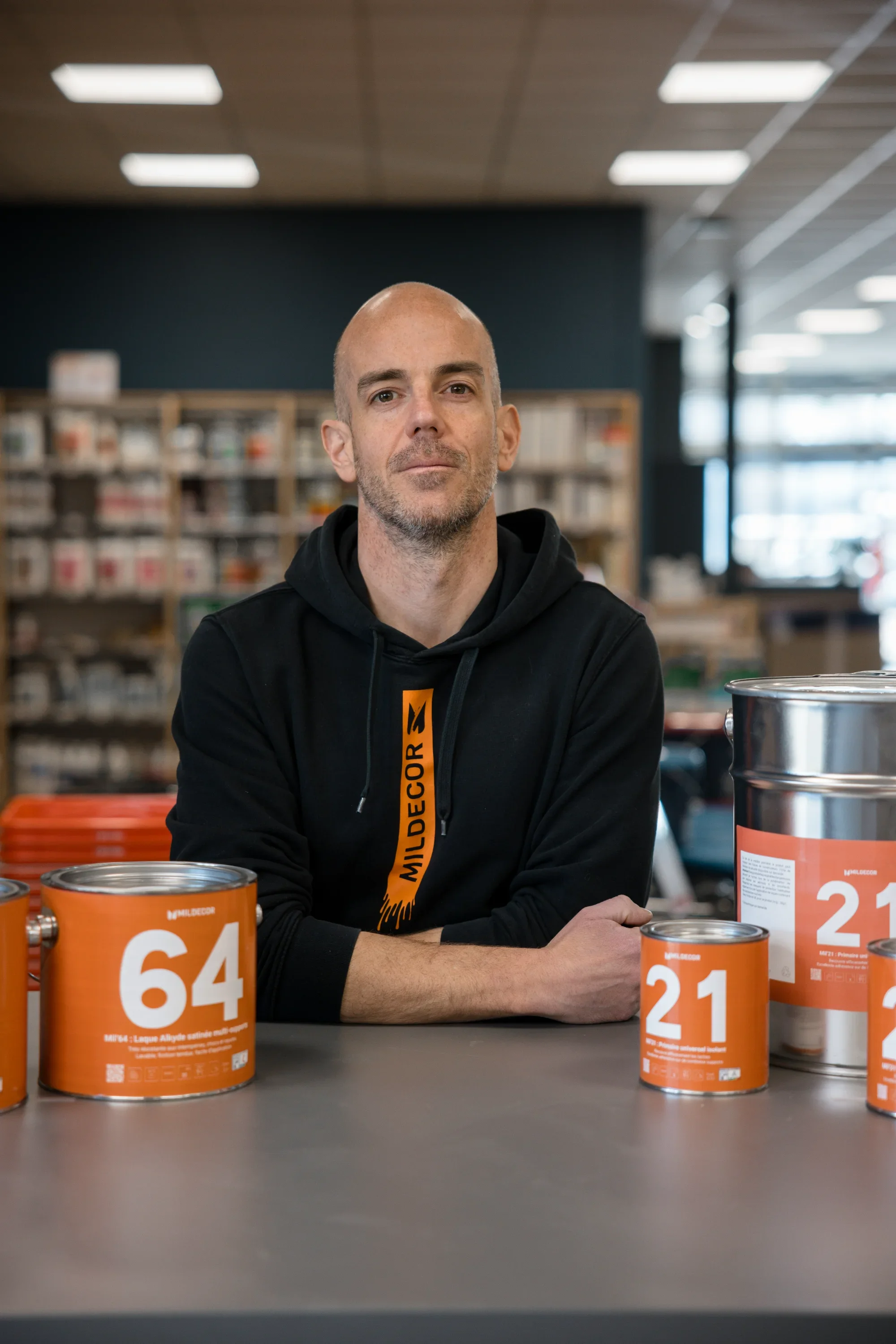 Un homme debout derrière un comptoir, à côté de plusieurs boîtes de peinture orange, dans un magasin.