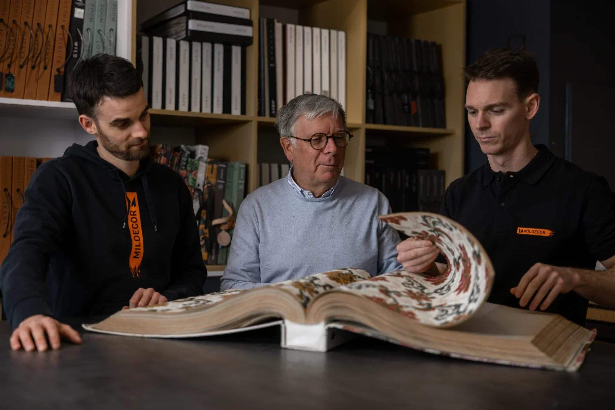 Trois hommes regardent un grand livre ou un tapisseries anciennes dans une bibliothèque.