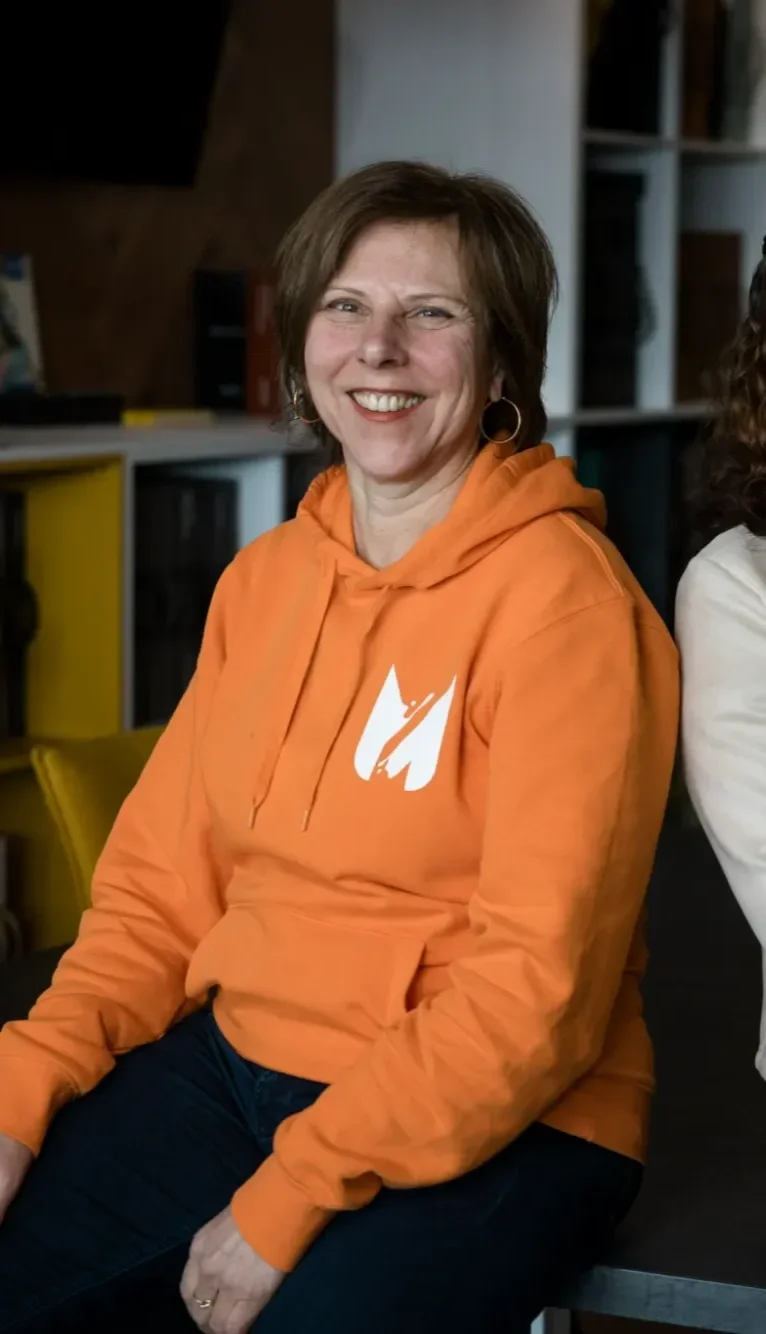 Femme souriante portant un sweat à capuche orange avec un logo blanc, assise dans un intérieur moderne.