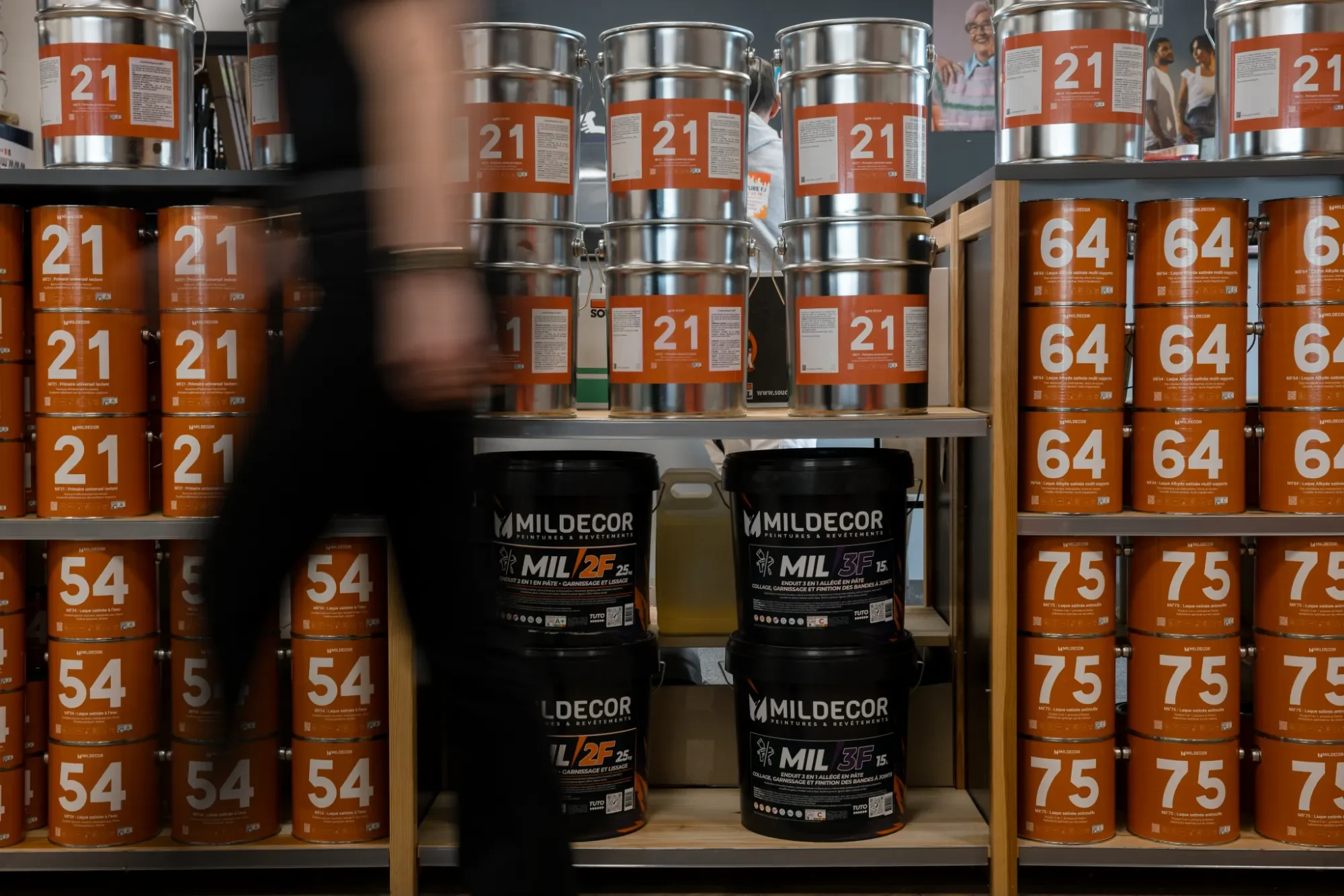 Rayons de pots de peinture orange et noir dans un magasin, avec des étagères en bois et des clients en arrière-plan.