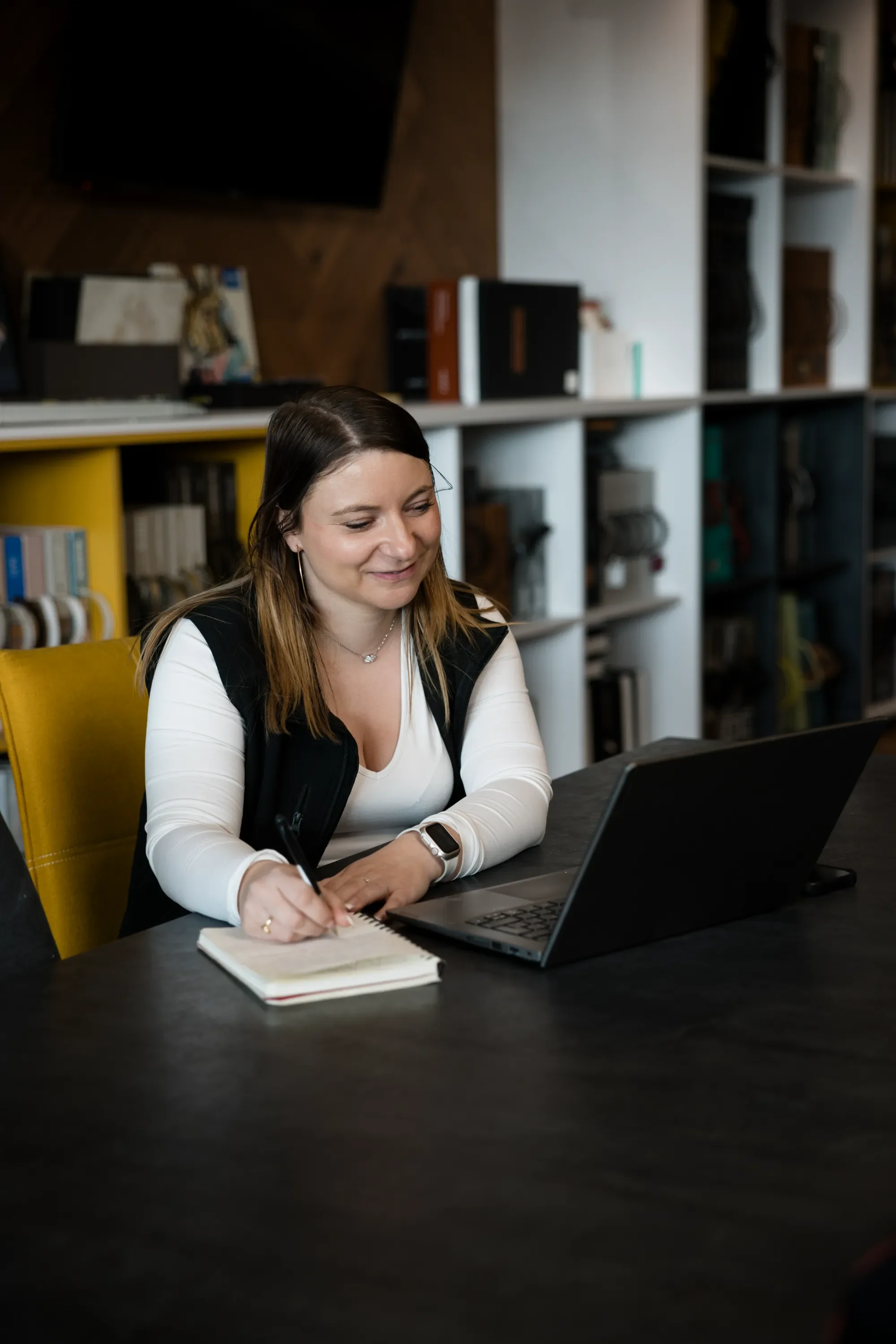Une femme assise à une table noire avec un ordinateur portable, prenant des notes dans un carnet, dans un bureau moderne avec étagères blanches remplies de livres et objets