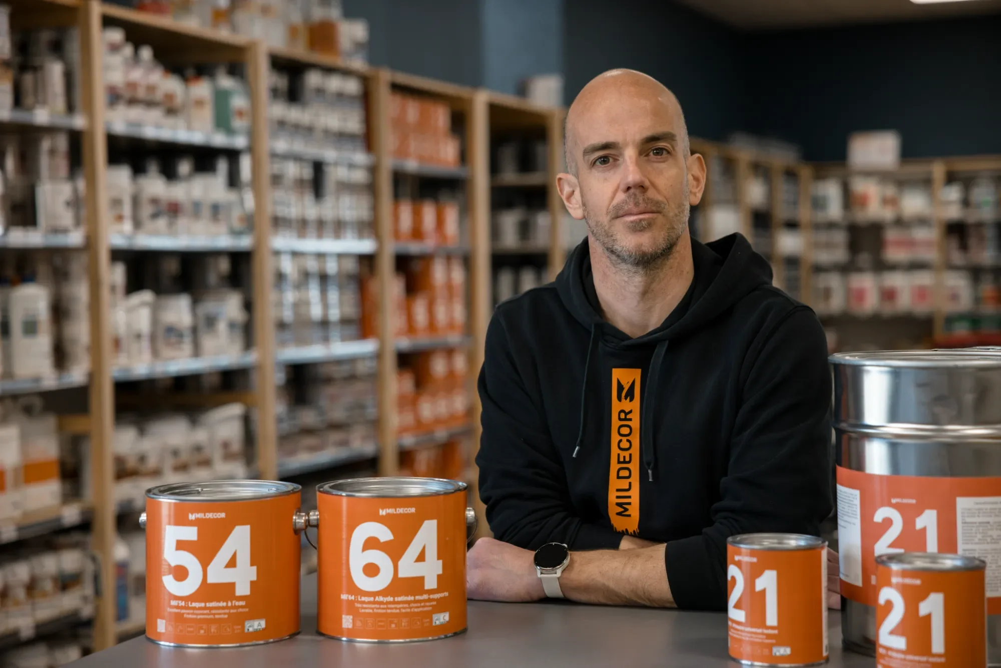 Un homme assis à une table dans un magasin de peinture, portant un sweat noir avec une étiquette orange inscrite "Mildecor". Devant lui, plusieurs pots de peinture orange avec des numéros comme 54, 64, et 21. En arrière-plan, des étagères remplies de