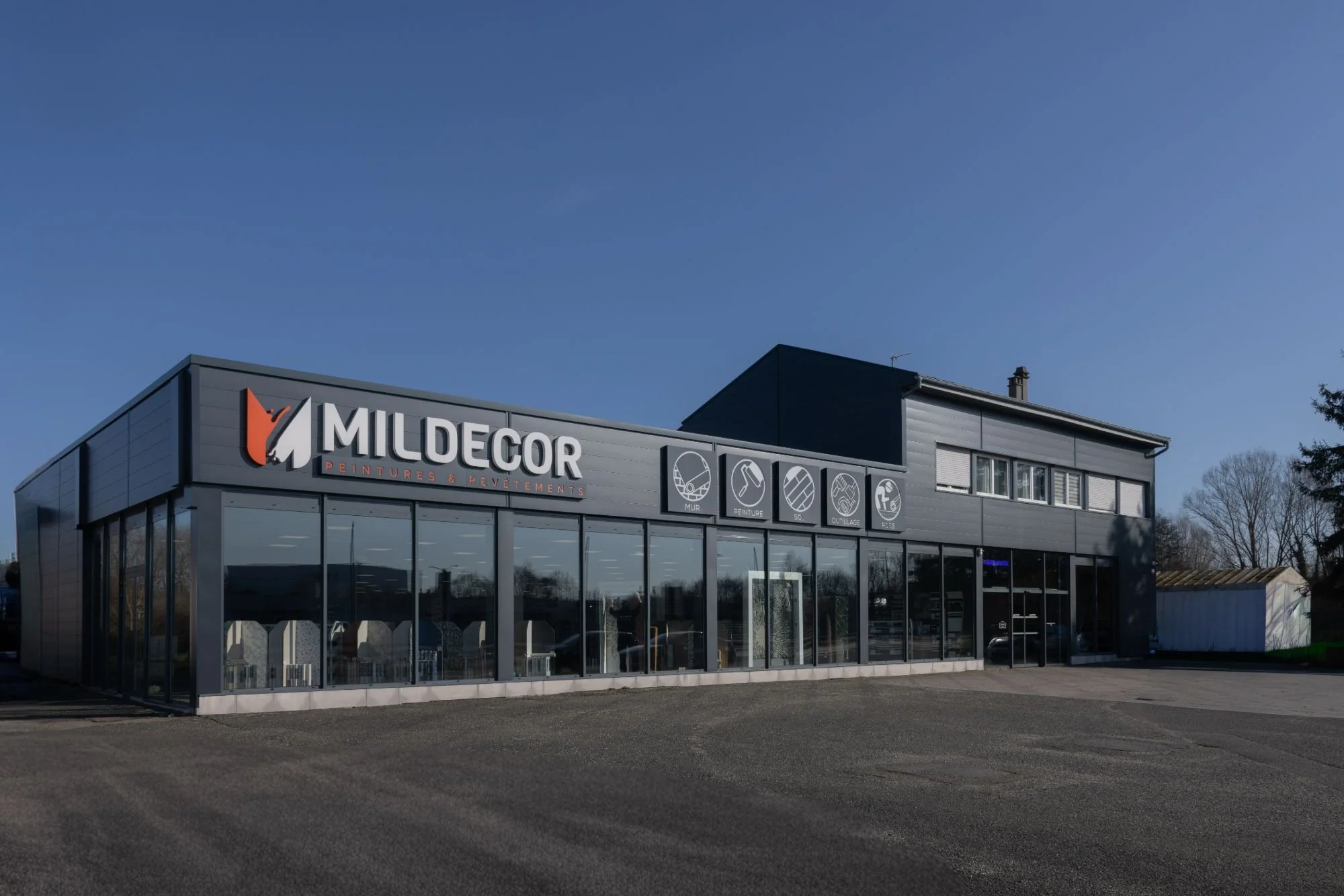 Commerce spécialisé en peintures et revêtements, nommé Mildecor, avec un bâtiment moderne en gros plan, comprenant un panneau d'enseigne et des icônes illustrant les services tels que mur, peinture, sol, cloison et façade, situé dans un environnement urbain avec un ciel clair.