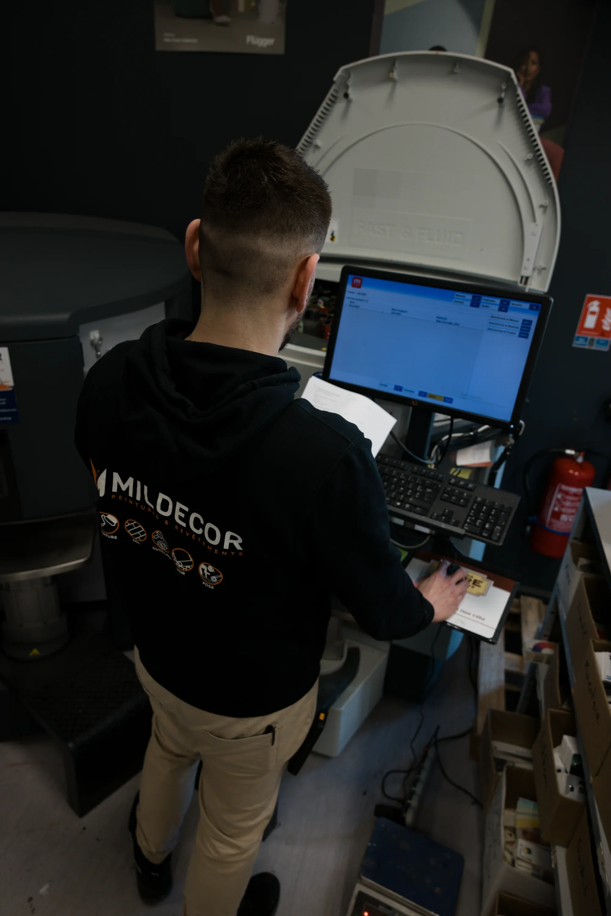 Une personne vue de dos utilise un ordinateur à une station de récupération d'argent dans un centre commercial ou un magasin. La personne porte un sweat à capuche noir avec le logo 'Mildecor' et des pictogrammes.