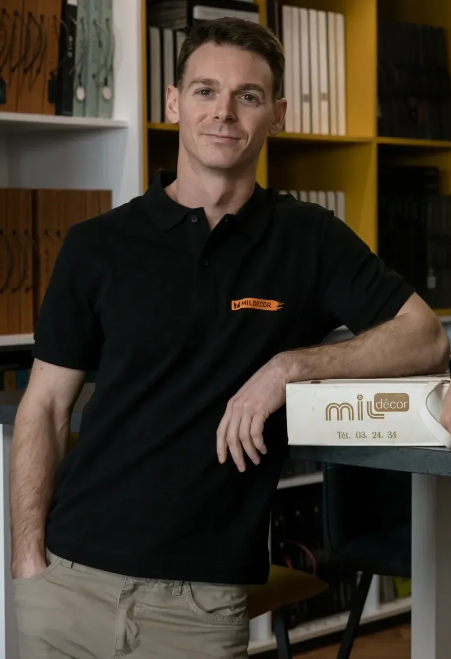 Un homme avec une chemise noire et des vêtements beige, posant dans une salle avec des étagères contenant des livres et des boîtes, avec un sourire léger.