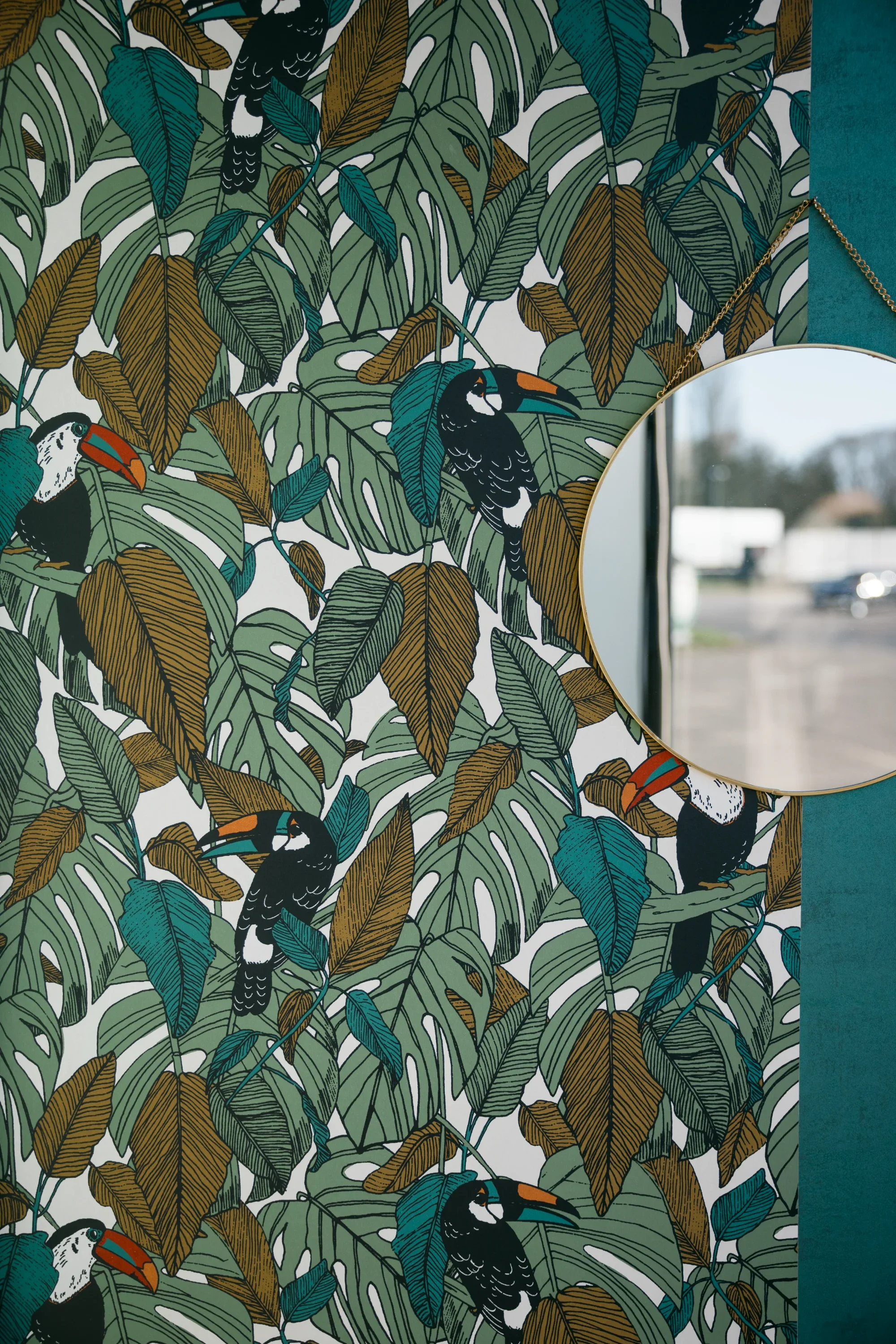 Murale à motif de feuilles tropicales avec toucans, partiellement visible dans un miroir rond suspendu sur le mur.