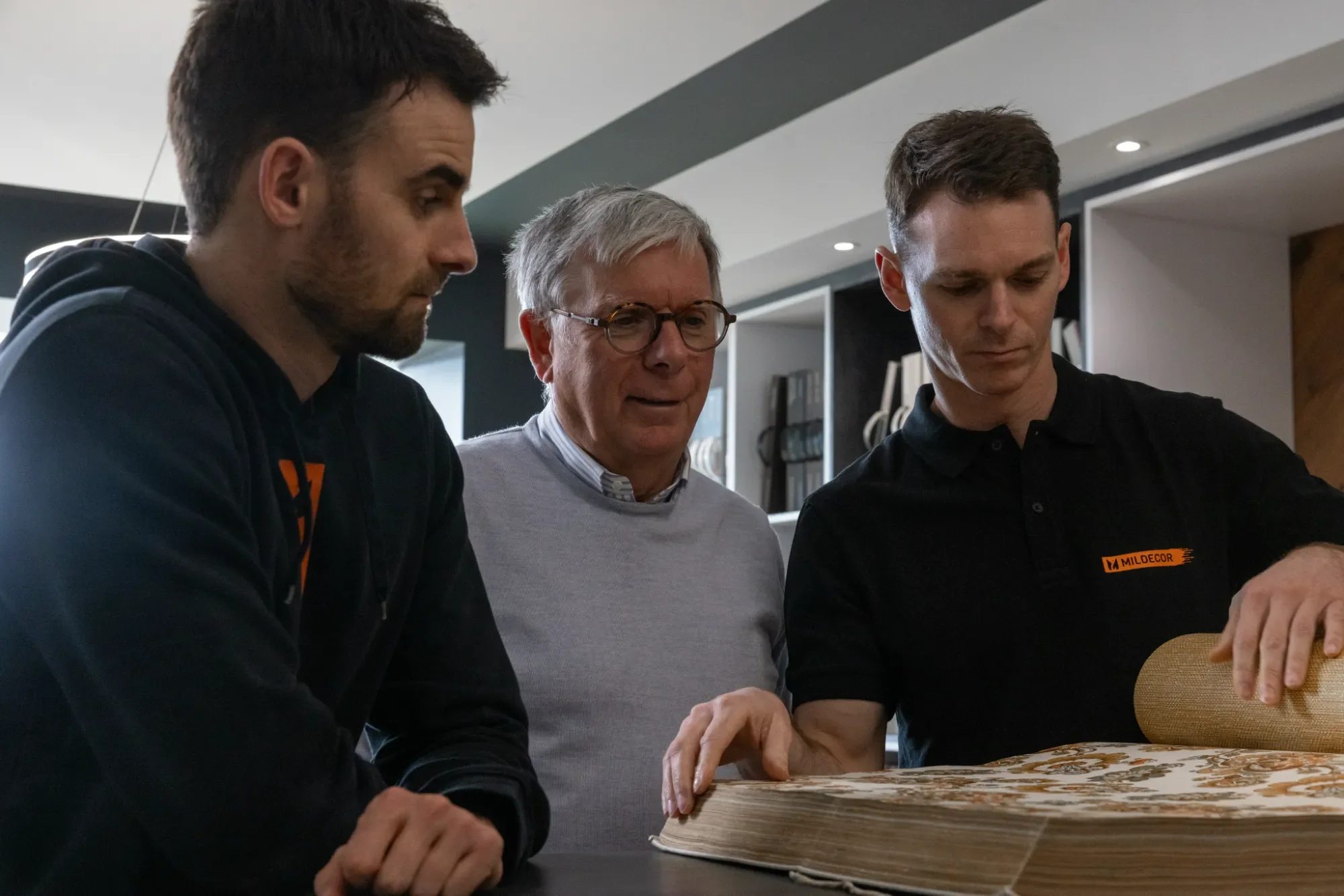 Trois hommes regardent un grand livre ou un échantillon de tissu sur une table dans un intérieur moderne.