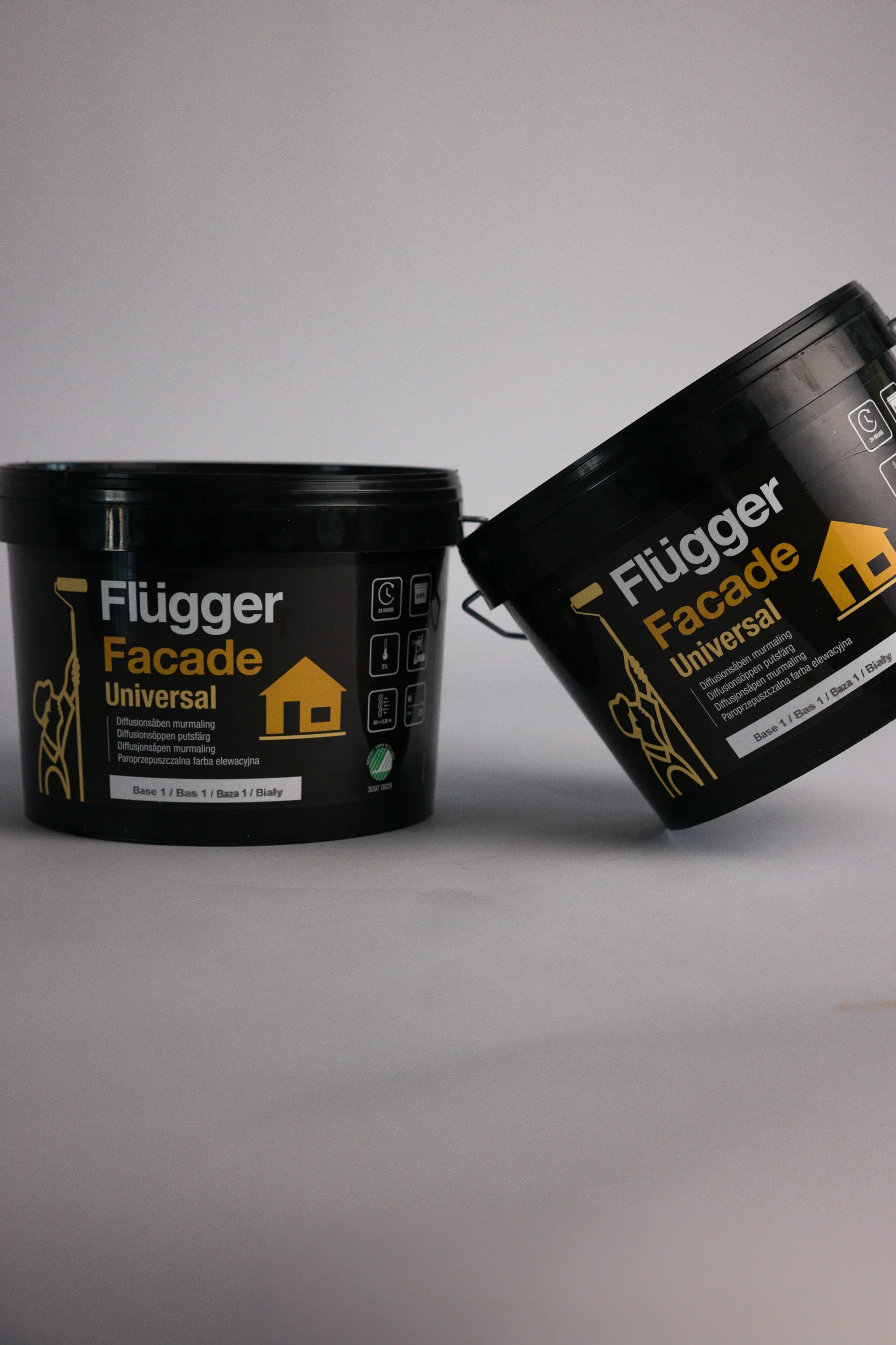 Deux bidons noirs de peinture ou de produit pour façade, l'un à gauche et l'autre à droite, avec des étiquettes noires et jaunes portant le texte "Flügger Facade Universal".