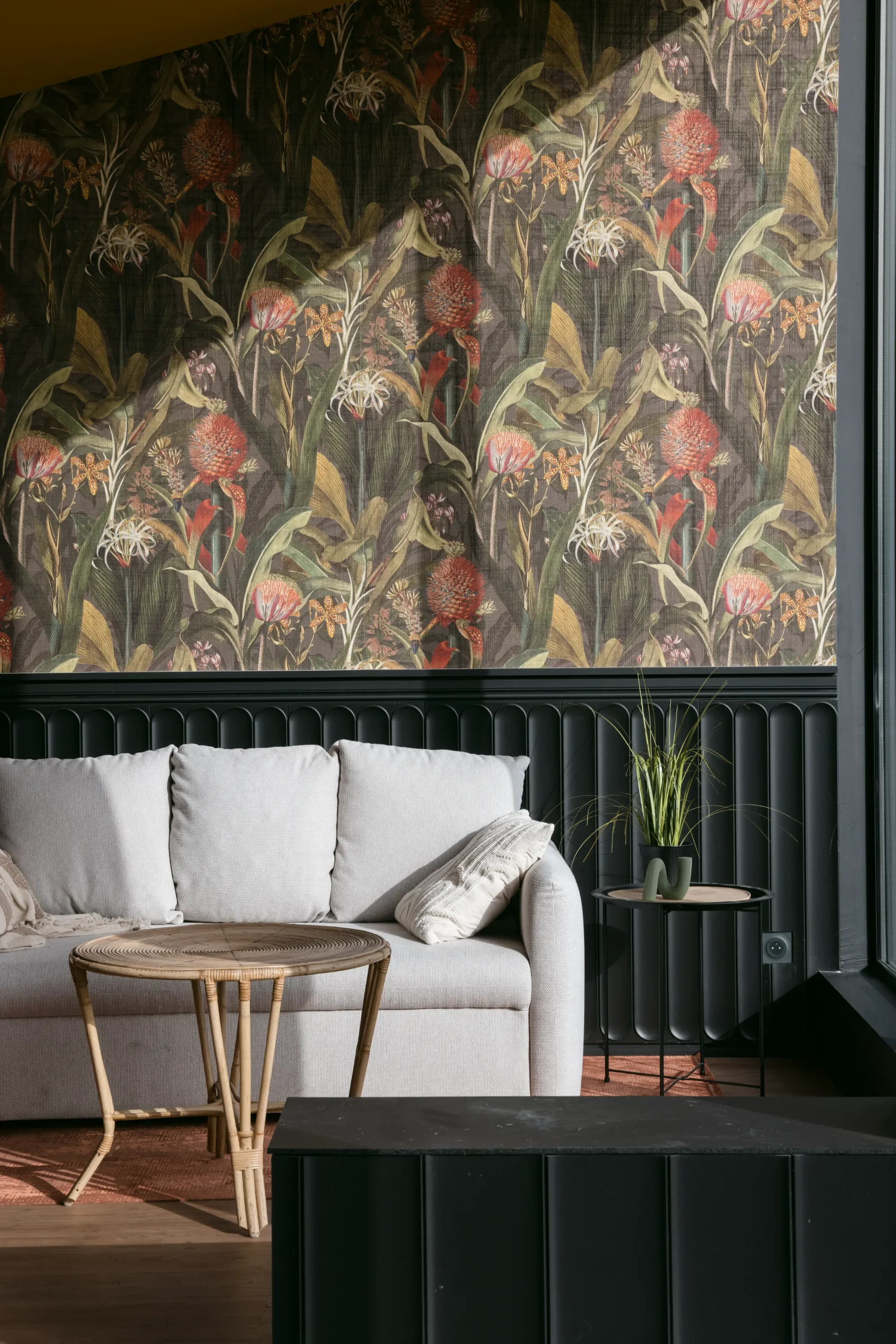 Intérieur d'un salon moderne avec un canapé beige, une table en bois, une plante verte sur une petite table noire, un mur tapissé de papier peint floral et une fenêtre.