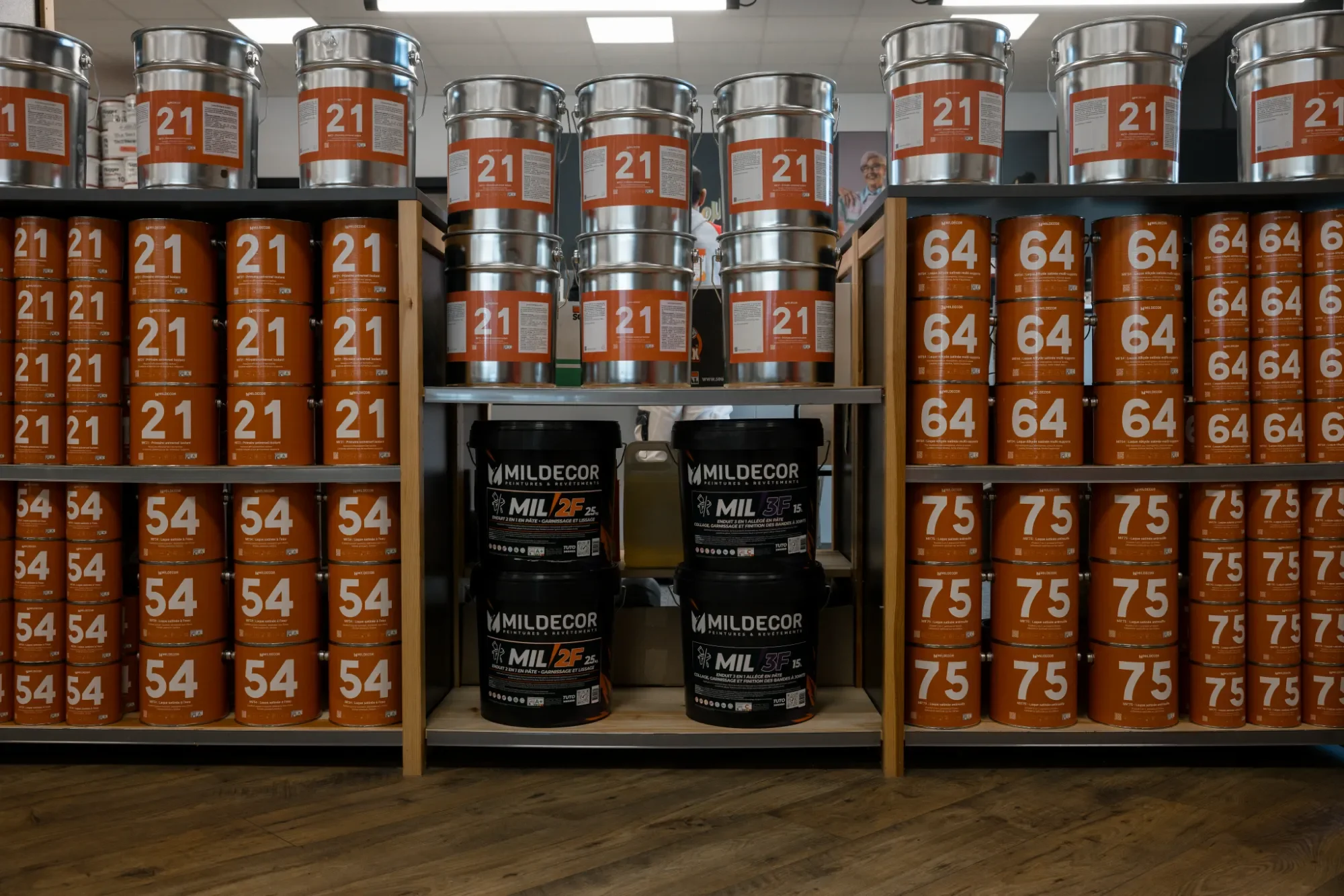 Étagères remplies de bidons de peinture orange et gris, avec deux seaux de peinture noirs au centre.