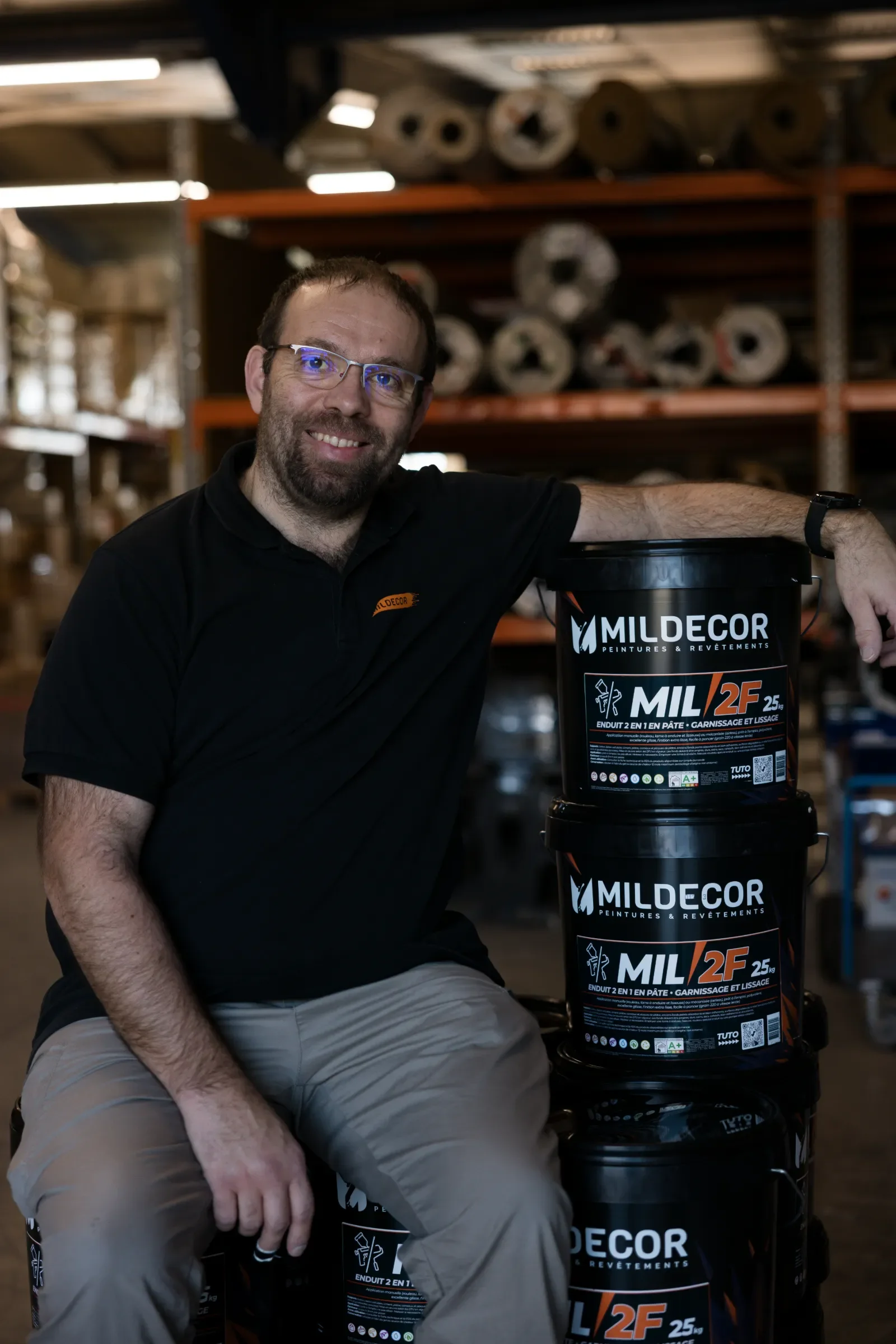 Un homme souriant, portant des lunettes, assis à côté de plusieurs bidons de peinture MILDECOR dans un entrepôt. Il porte un polo noir avec un logo orange, derrière des étagères remplies de rouleaux de papier ou de matériaux.