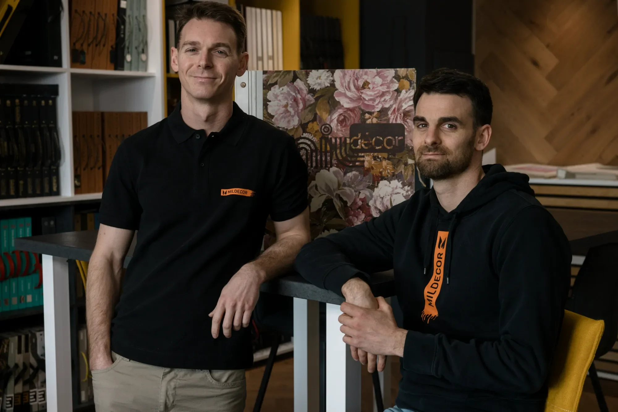 Deux hommes posent dans un intérieur moderne, portant tous deux des vêtements noirs avec le logo Mildecor, devant une bibliothèque avec des livres et un décor floral derrière eux.