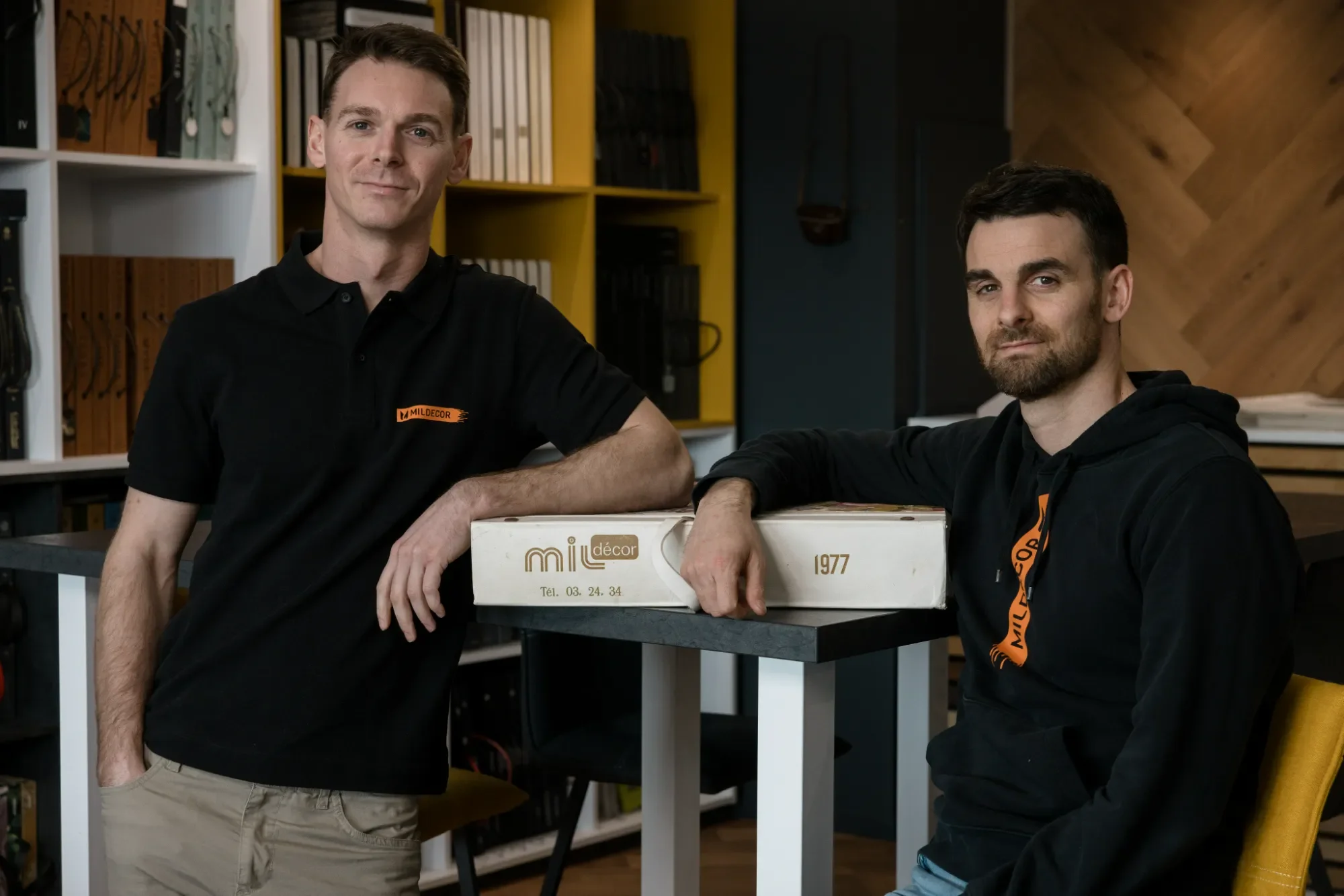 Deux hommes posant avec une boîte à côté d'une étagère remplie de livres et d'objets, dans un intérieur moderne.