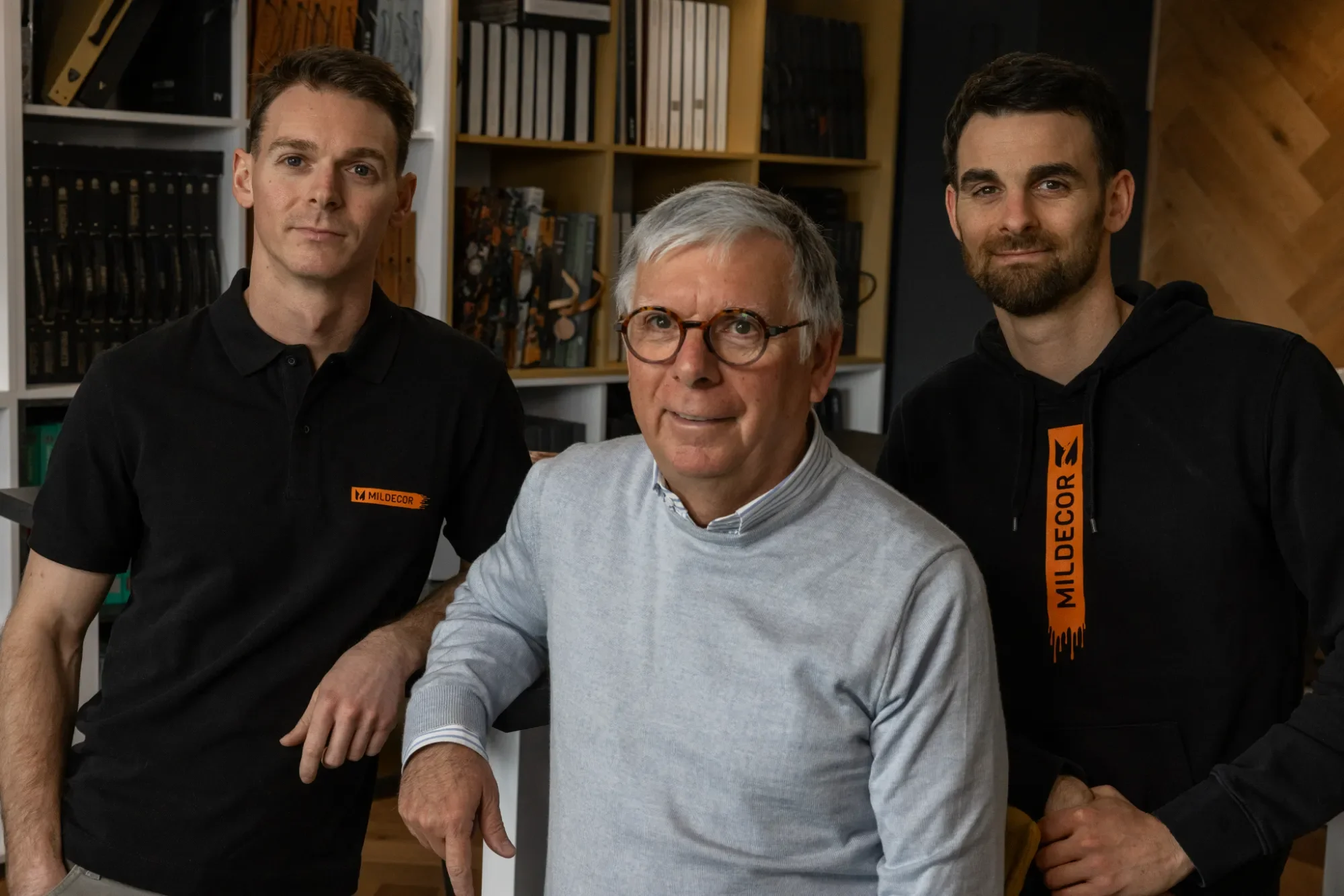 Trois hommes posant ensemble dans un bureau, deux portent des sweat-shirts noirs avec le logo MILD E COR, un homme en pull gris clair, arrière-plan avec étagères remplies de livres et de boîtes.