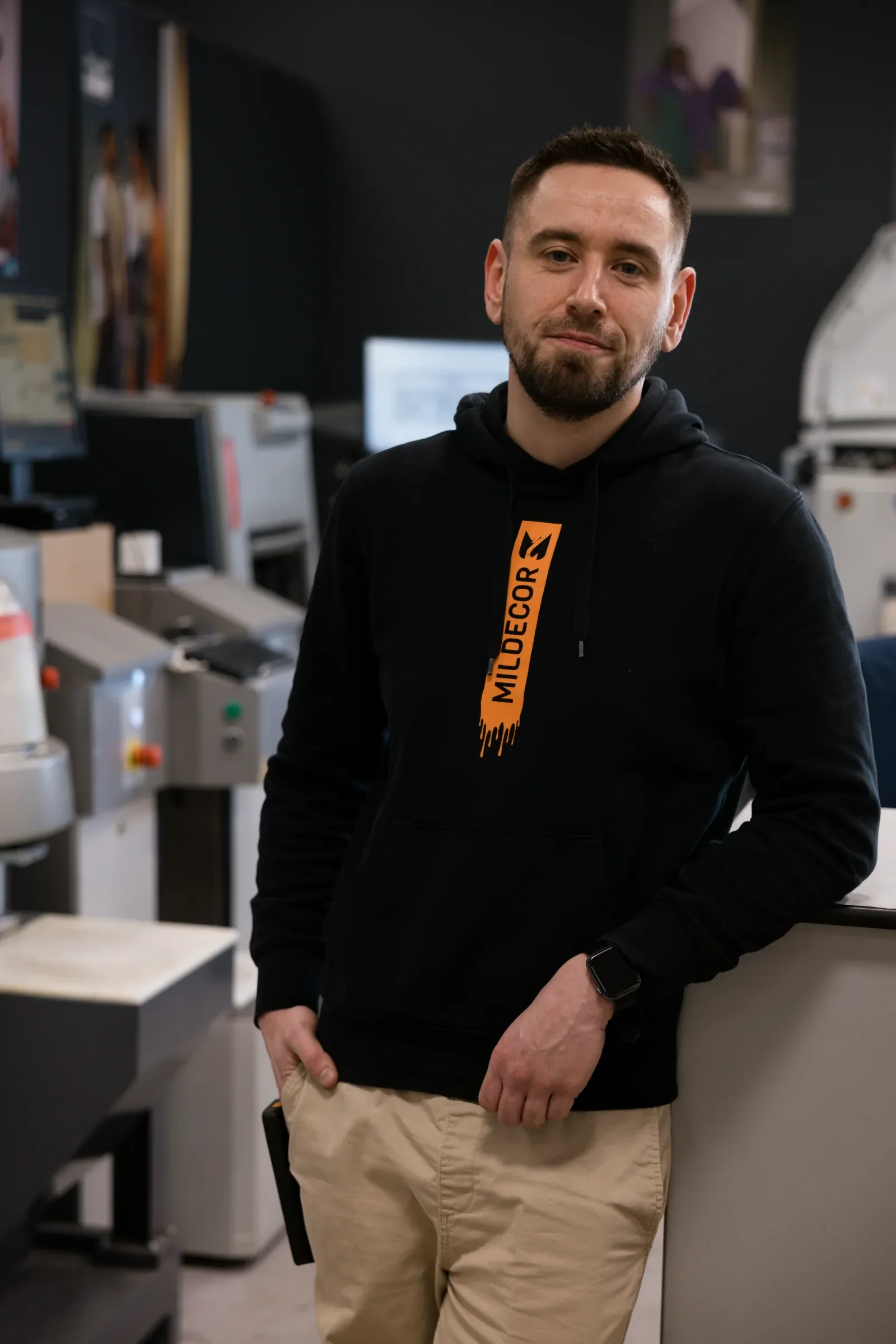 Jeune homme avec une barbe, portant un sweat noir avec un logo orange, et un smartwatch noir, debout dans un environnement de bureau ou de laboratoire avec des ordinateurs et des machines en arrière-plan.
