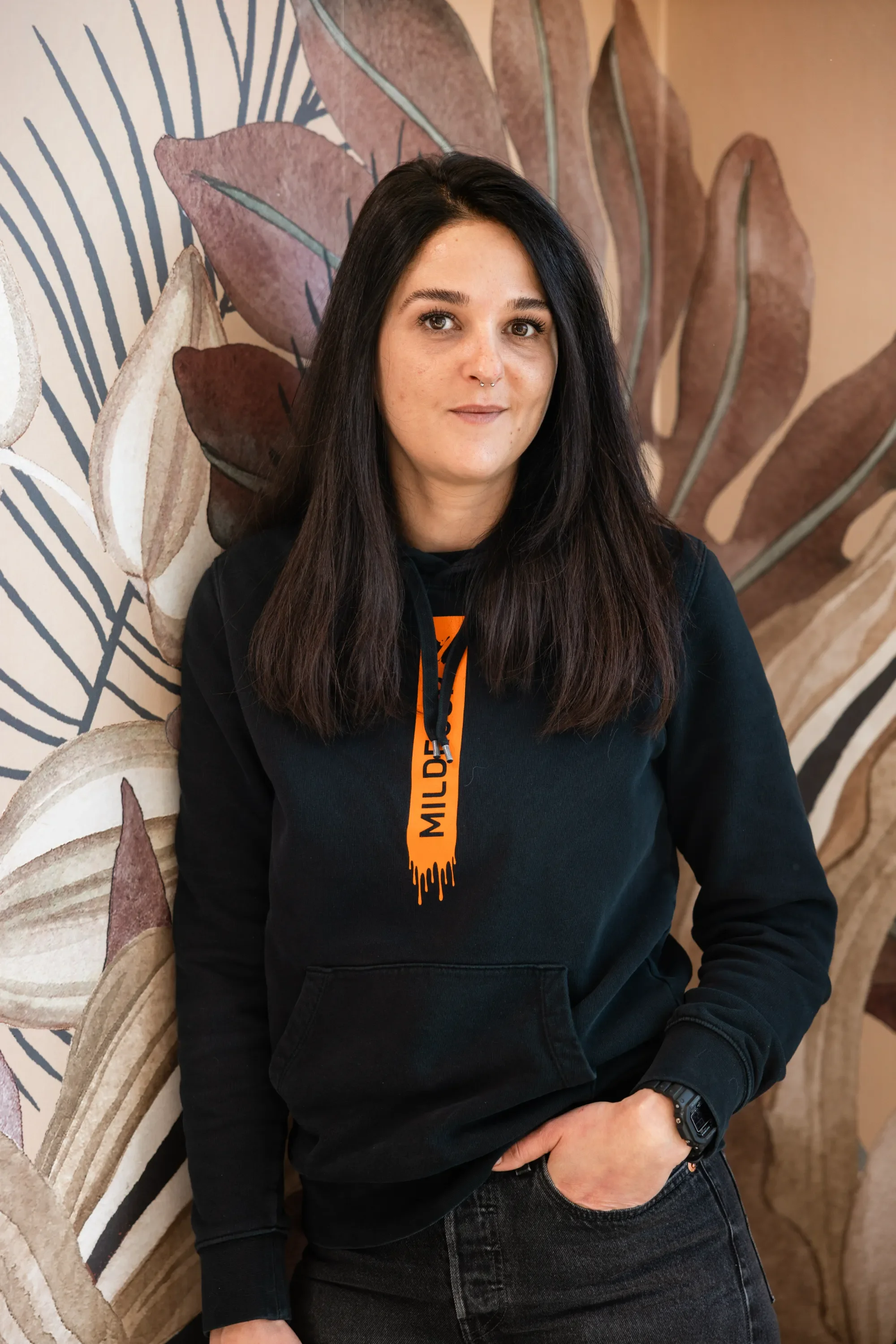 Jeune femme aux cheveux noirs, portant un sweat noir avec un logo orange, debout devant un mur avec un motif de feuilles en beige et rose.