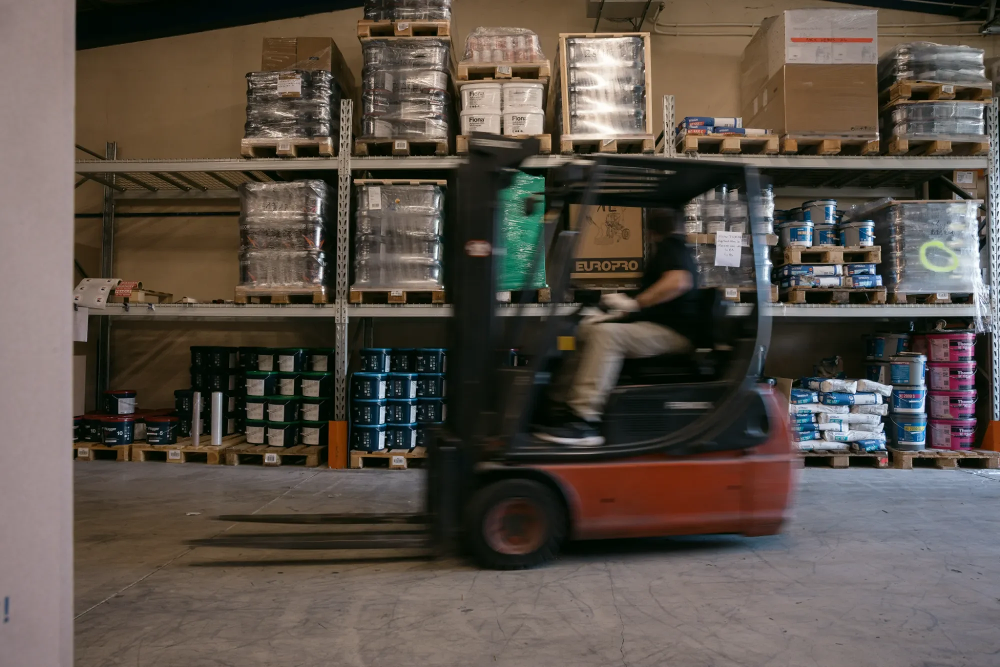 Un élévateur roulant transporte des palettes de matériaux stockés dans un entrepôt. En arrière-plan, des étagères remplies de caisses, de bidons et d'emballages en plastique.