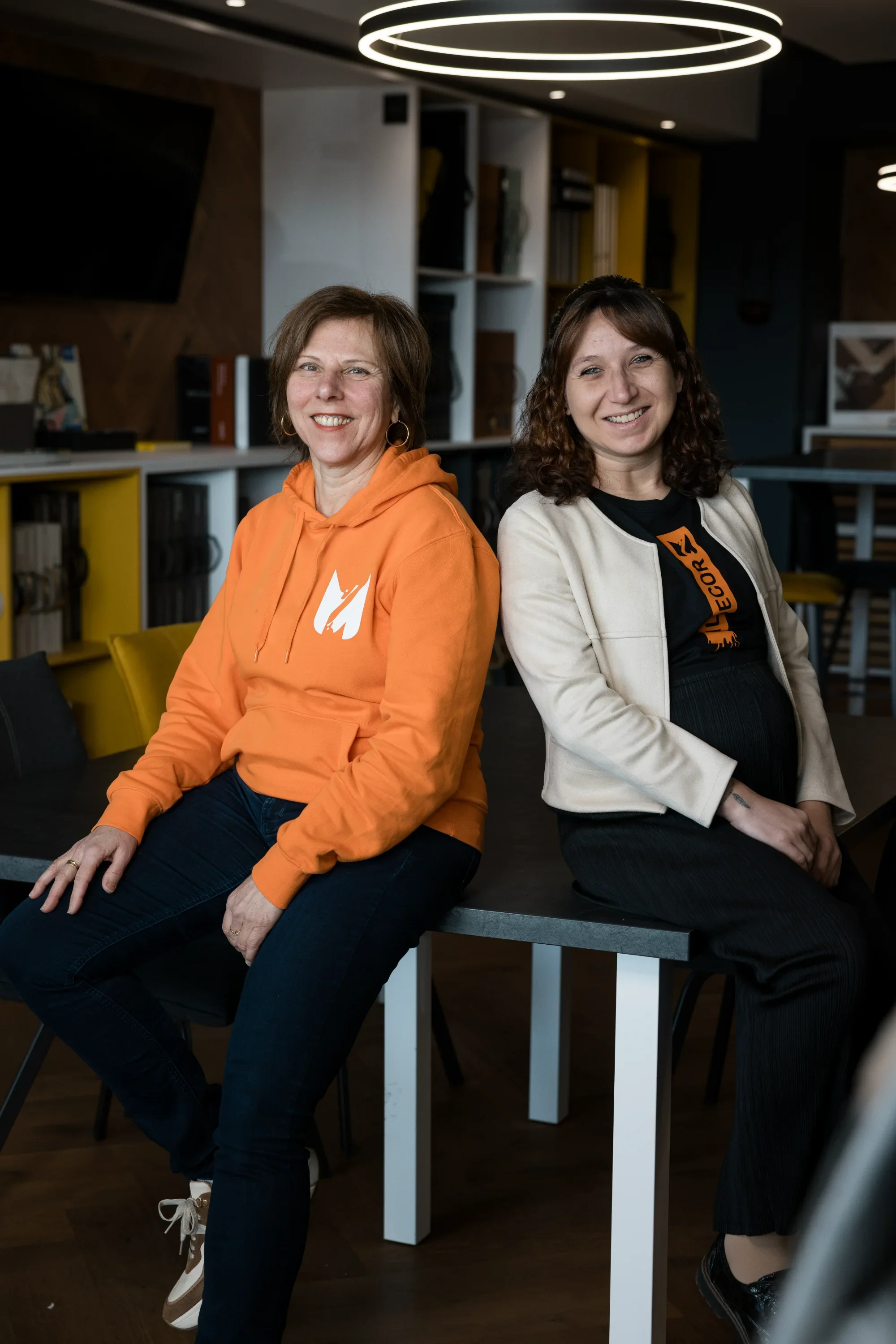 Deux femmes assises sur une table dans un espace moderne, souriant à la caméra. L'une porte un sweat orange, l'autre une veste beige.
