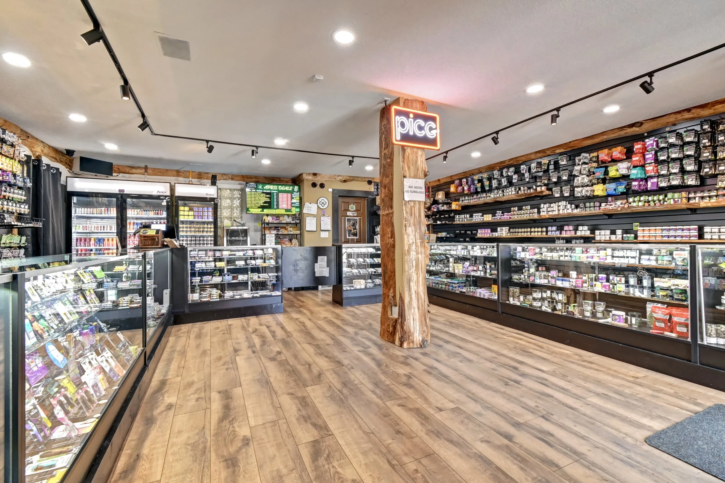 Interior of a retail store with glass display cases filled with products, shelves stocked with various items, wood flooring, bright ceiling lights, and a neon sign that reads 'picc' on a wooden support beam.