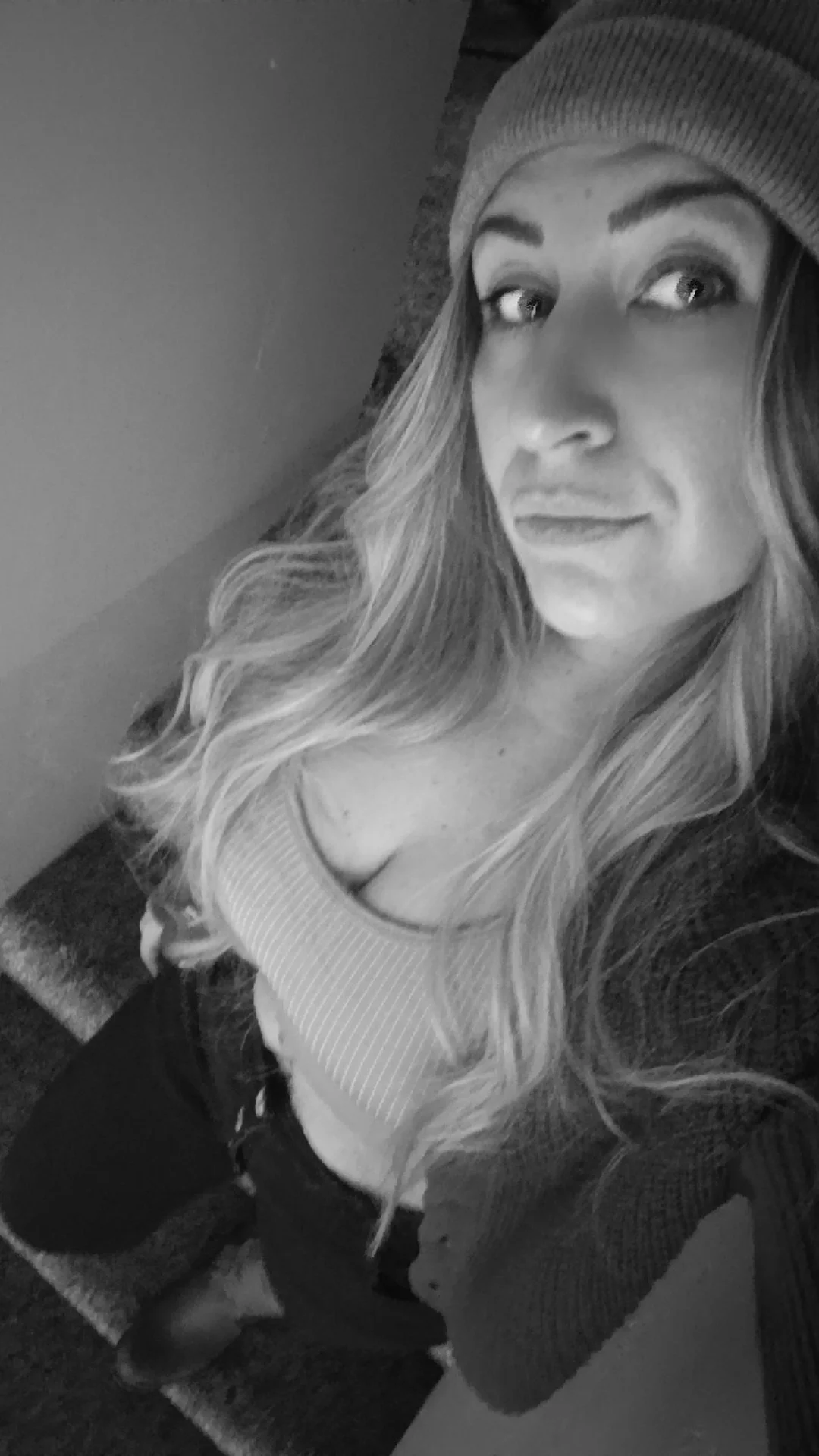 Black and white selfie of a woman with long wavy hair, wearing a beanie, taken from a high angle, showing her smiling face and part of her body.