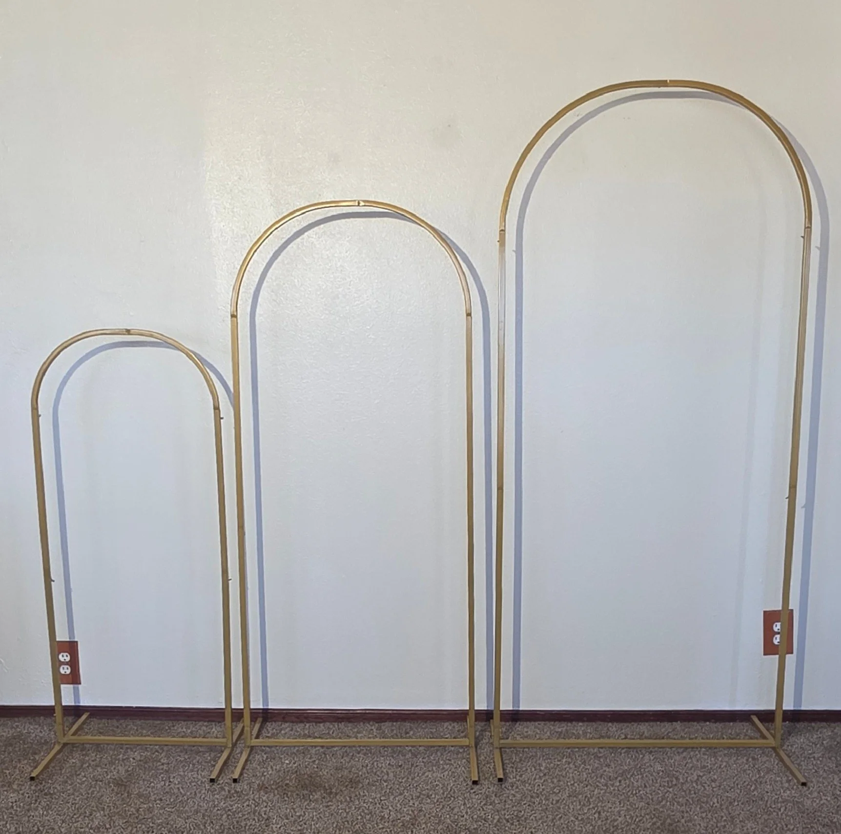 Three empty clothing racks with arch-shaped frames in various sizes against a plain white wall, with electrical outlets visible at the bottom.