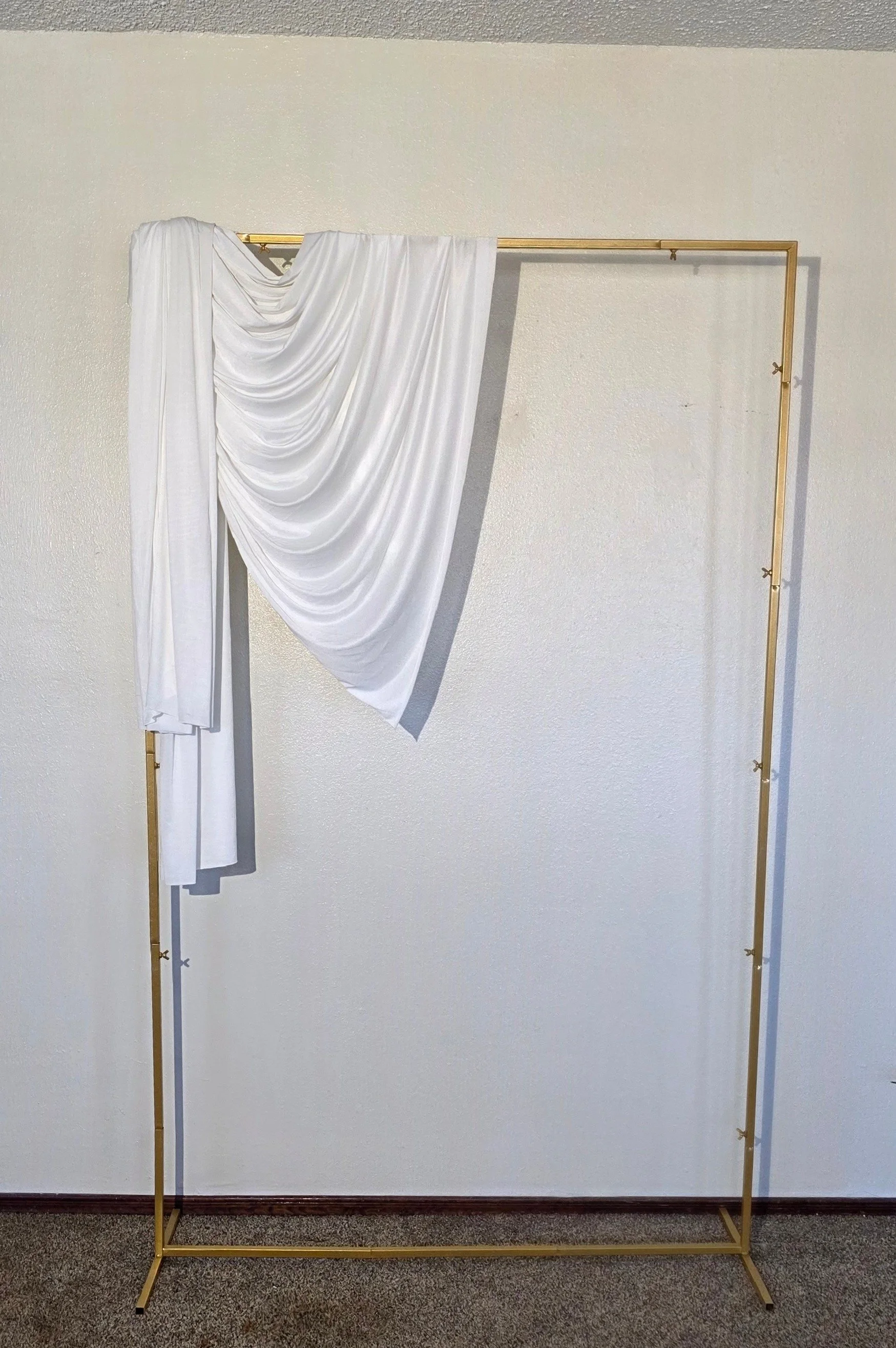 A gold-colored clothing rack with white drapes hanging on it, partially covering the open frame, placed against a white wall.