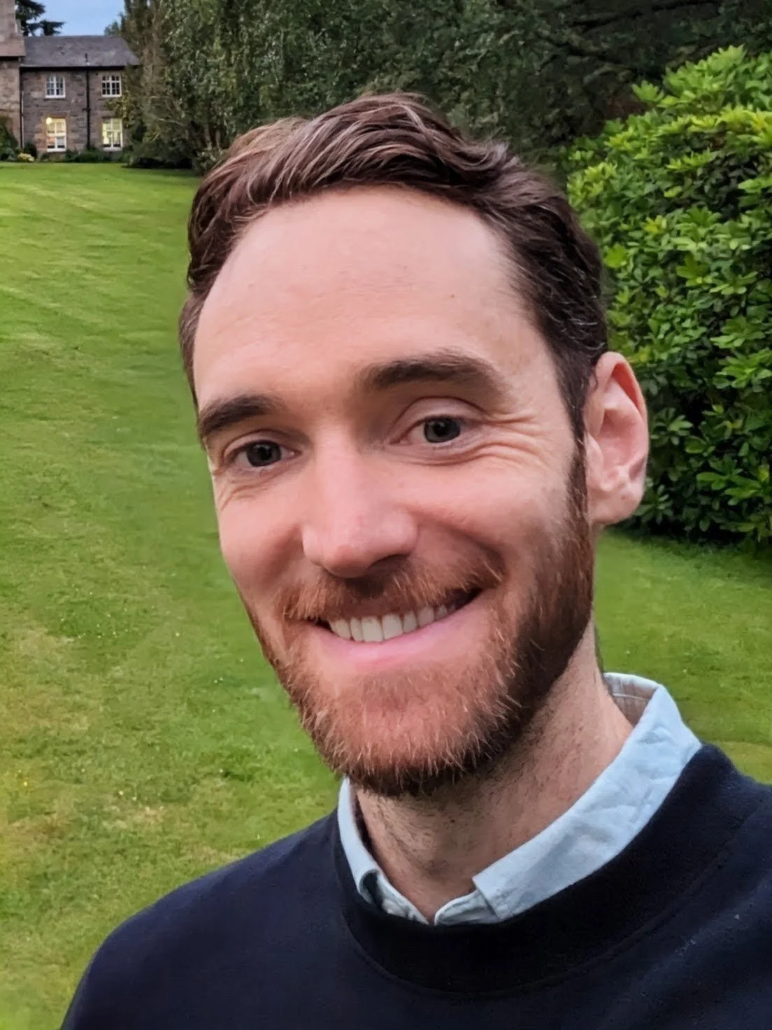 A smiling man with a beard and short brown hair, wearing a collared shirt and a dark sweater, standing outdoors in a lush green yard with trees and bushes, and a large house in the background.