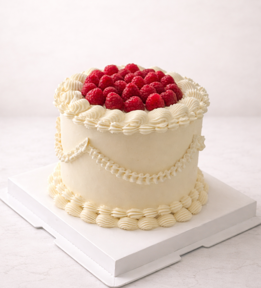 A white, heart-shaped cake decorated with raspberry fruit on top and cream swirls along the edges.