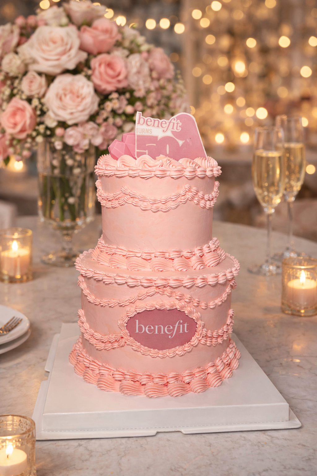 A pink, three-tiered Benefit Cosmetics themed birthday cake with pink decorative frosting and a topper that says 'Benefit turns 50.' The cake is on a white base, with a vase of pink roses and champagne glasses in the background, along with candles and out-of-focus warm lights.
