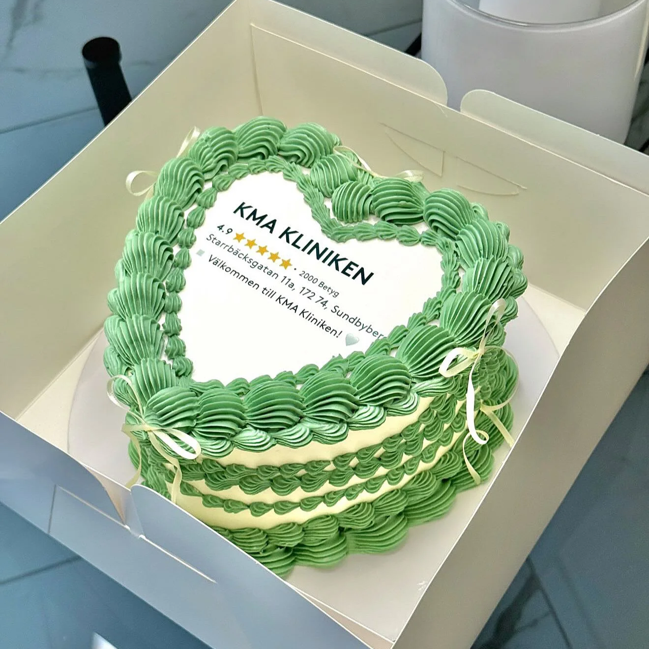 A heart-shaped cake with green ruffled icing border and white icing is inside a white box. The cake has a digital print with text in Swedish, indicating it is from KMA Kliniken, with a 4.9-star rating from 2000 reviews, and includes an address and greeting.