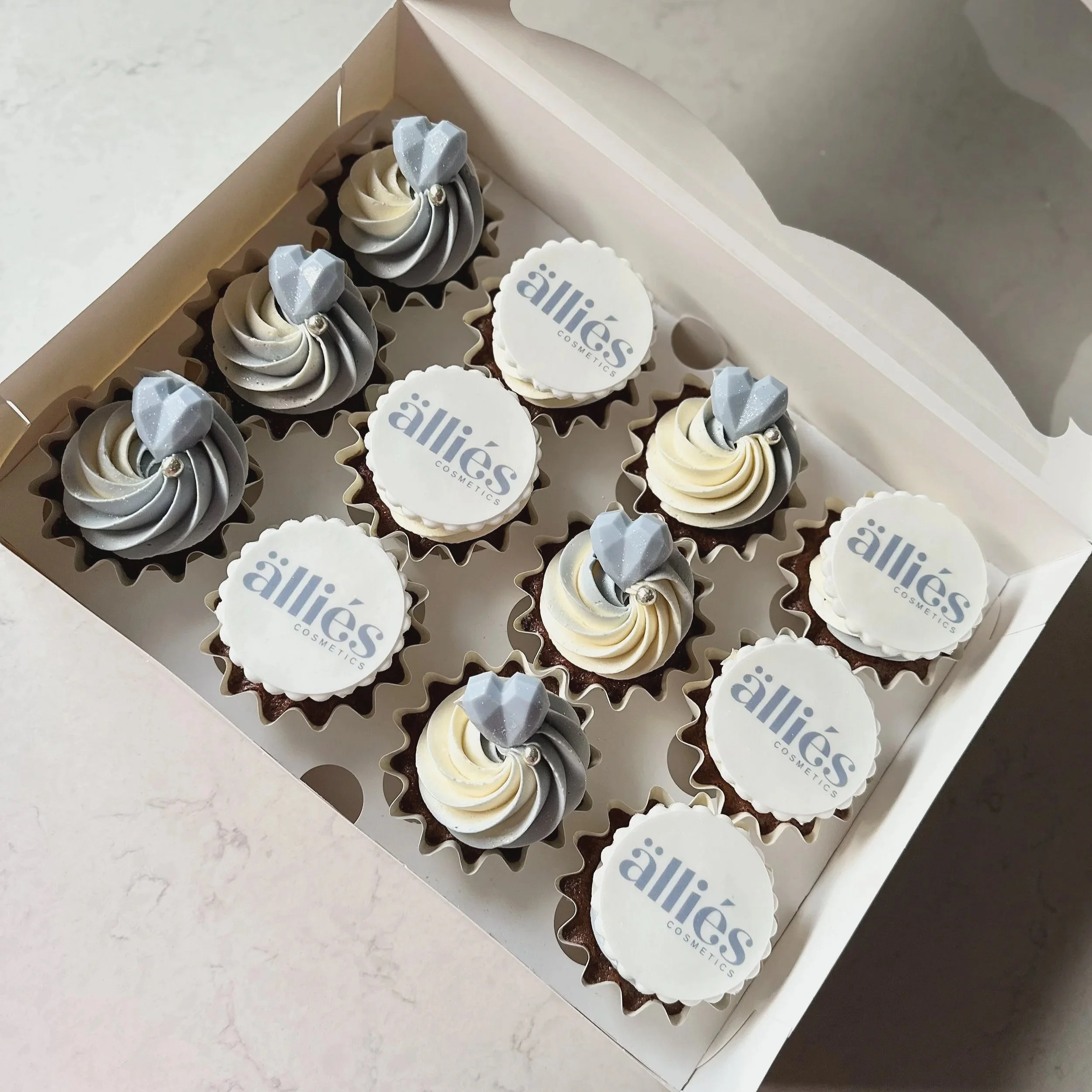 A white box containing ten cupcakes decorated with gray and white ice cream swirl frosting, each topped with a gray heart topper or a round edible logo reading 'allies cosmetics' in gray text.