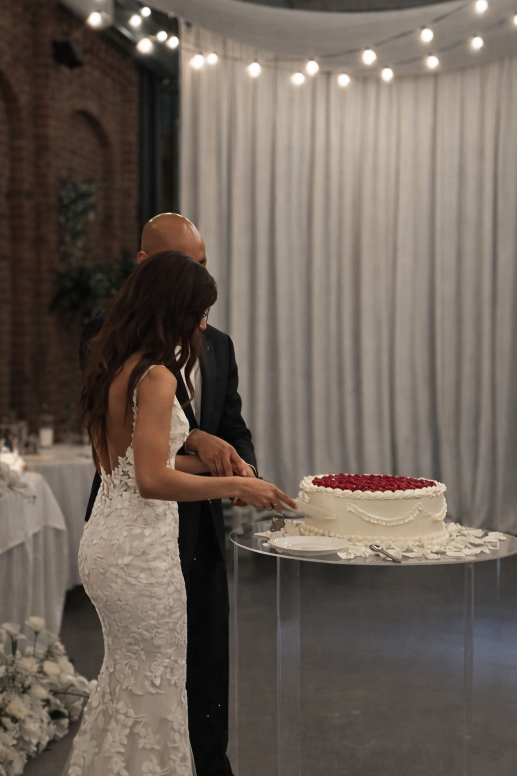 A bride and groom cutting a wedding cake together at their reception. The bride is wearing a white lace dress, and the groom is in a black suit. The cake is large, round, decorated with white icing and red fruit on top, and placed on a clear table with sugar rose petals around the base.