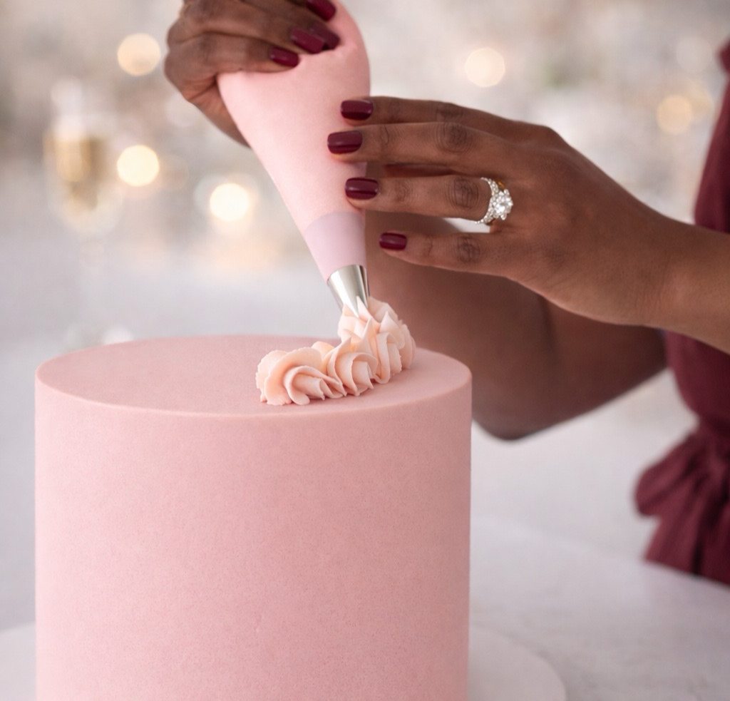 A person with dark skin and red nails decorates a pink cake with pink frosting using a piping bag.